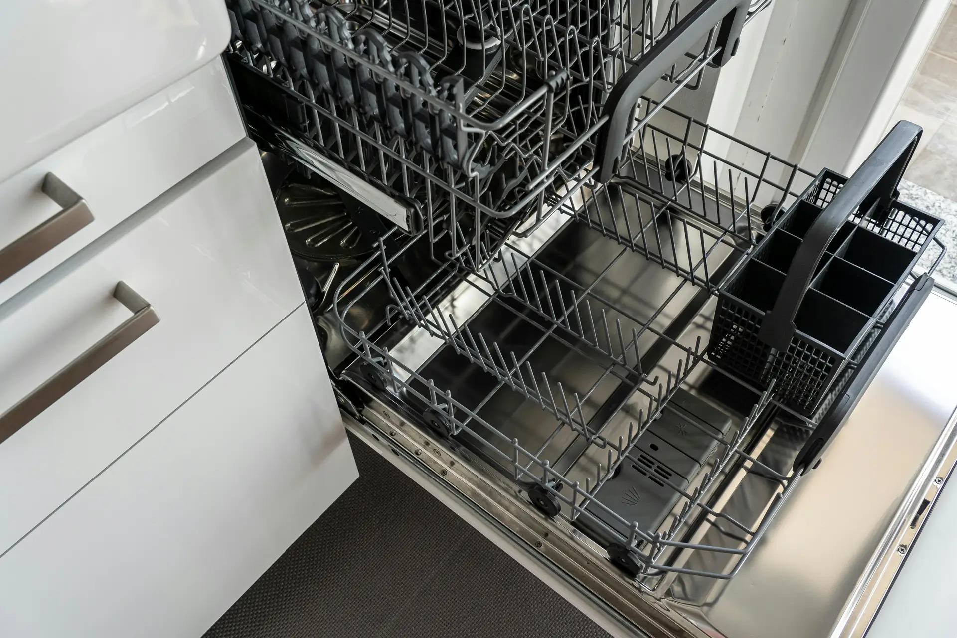 An open, empty dishwasher with pulled-out racks and a visible cutlery basket inside a modern kitchen, illustrating a spring cleaning checklist step focused on cleaning the dishwasher and maintaining household appliances.