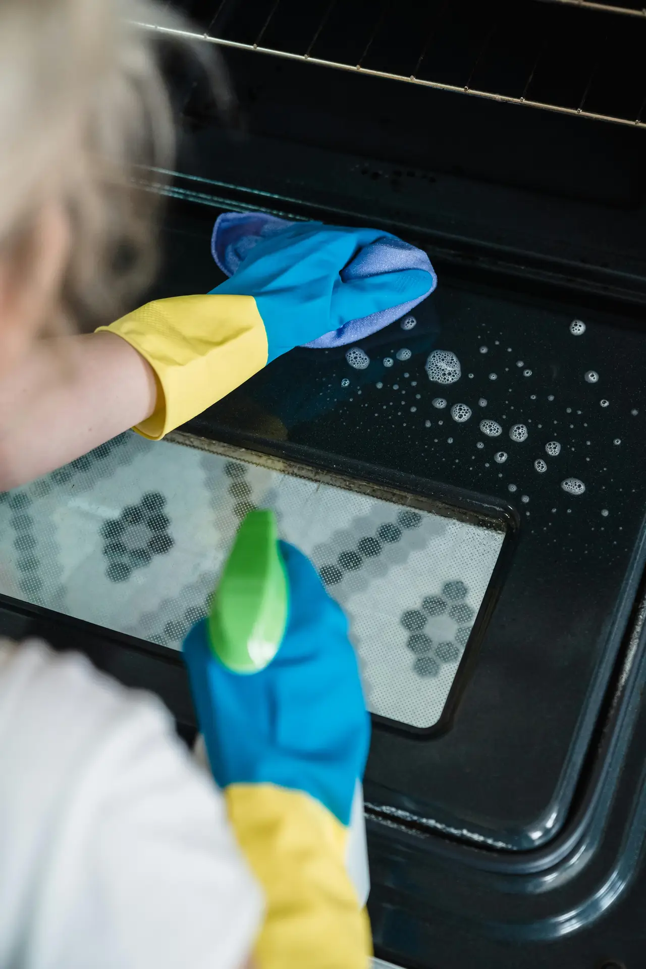 A person wearing protective gloves sprays cleaner and wipes the inside glass of an oven door, illustrating a spring cleaning checklist task focused on degrease the oven by removing baked-on residue and grease buildup.