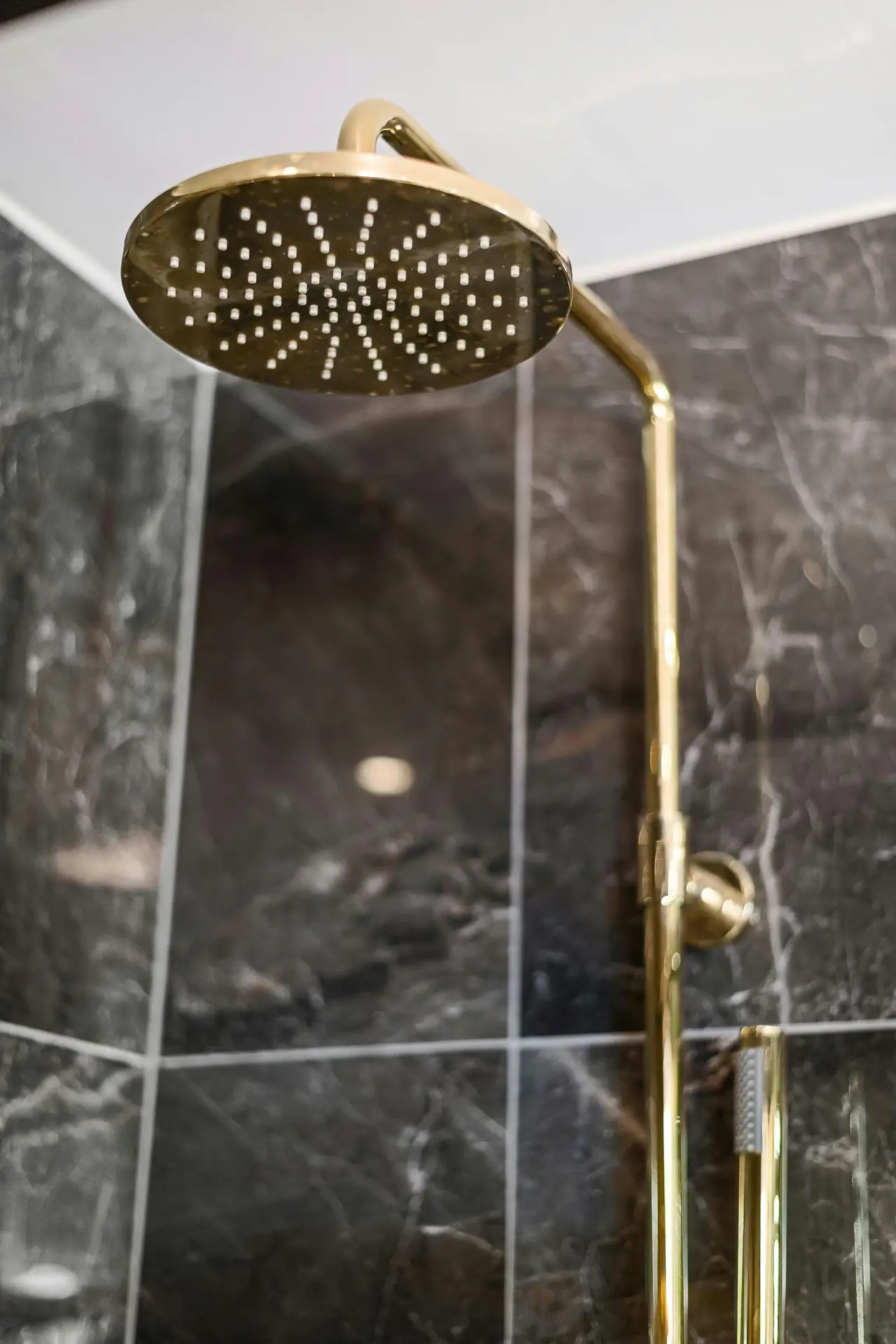 A close-up of a gold rain-style shower head mounted against dark stone tiles, illustrating a spring cleaning checklist task focused on descaling the shower head to improve water flow and maintain a fresh, well-kept bathroom.