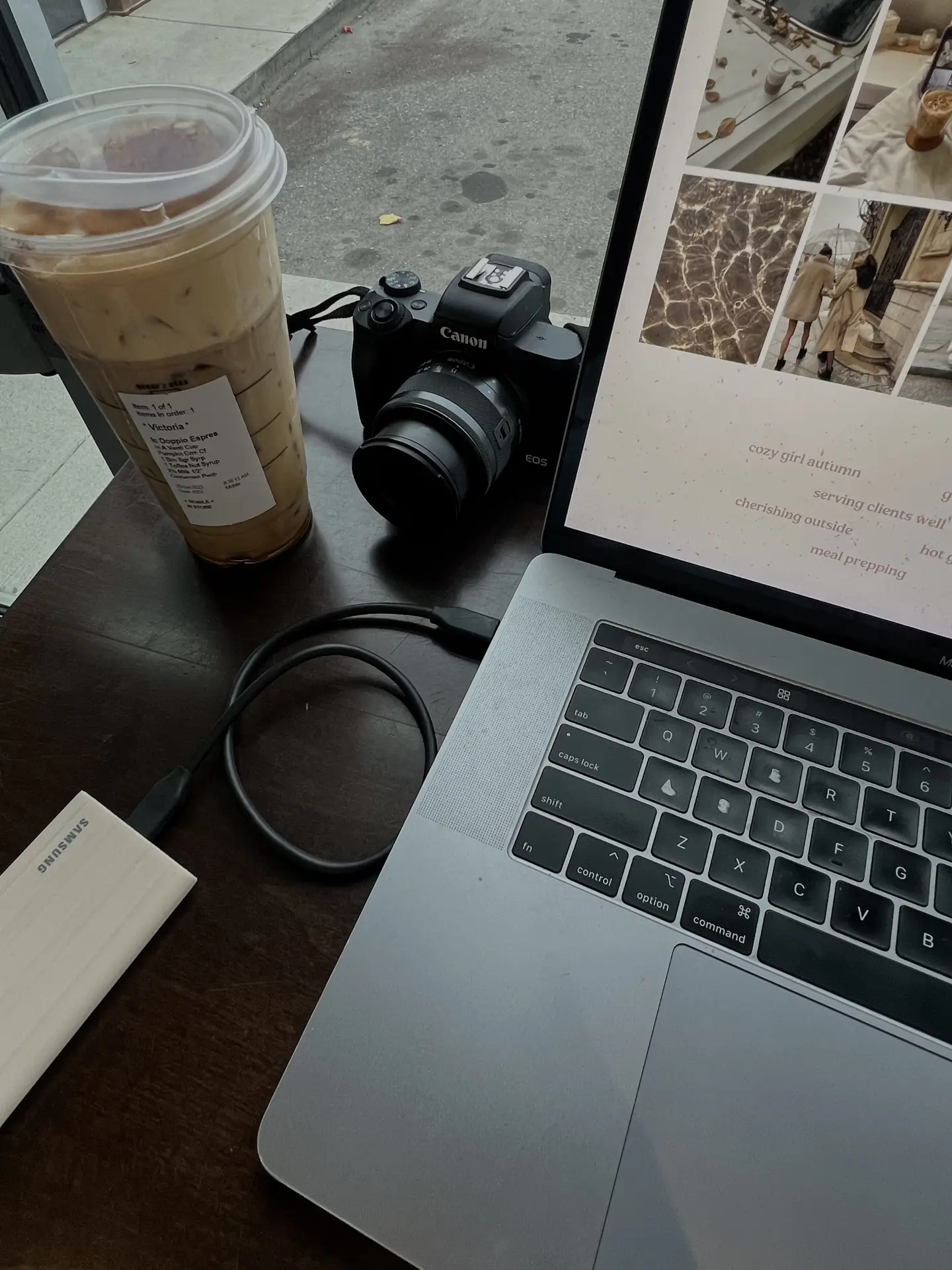 Laptop open on a café table next to a camera, iced coffee, and charging cable, illustrating a spring cleaning checklist moment focused on digital cleaning, organizing files, and streamlining creative work while on the go.