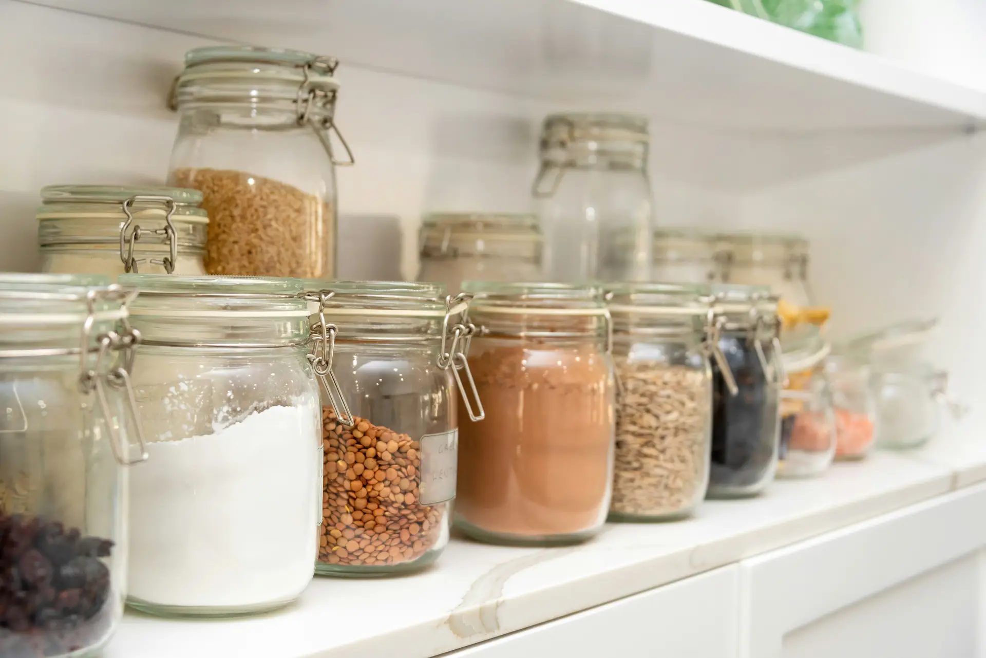 Neatly arranged glass jars filled with dry goods like flour, lentils, grains, and spices line a bright pantry shelf, representing a spring cleaning checklist task focused on organizing the pantry in a clean, minimalist kitchen setting.