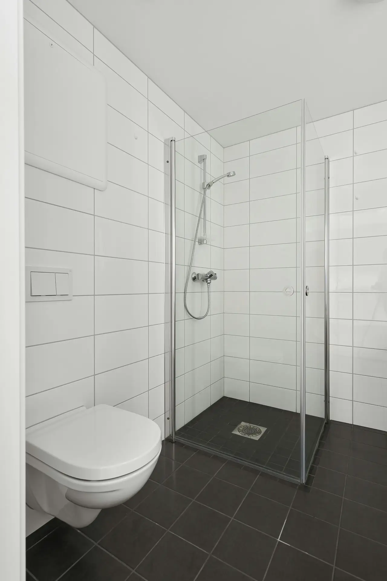 A minimalist bathroom with white wall tiles, dark floor tiles, a walk-in glass shower, and a wall-mounted toilet, representing a spring cleaning checklist task focused on refreshing grout to restore a clean and polished look.