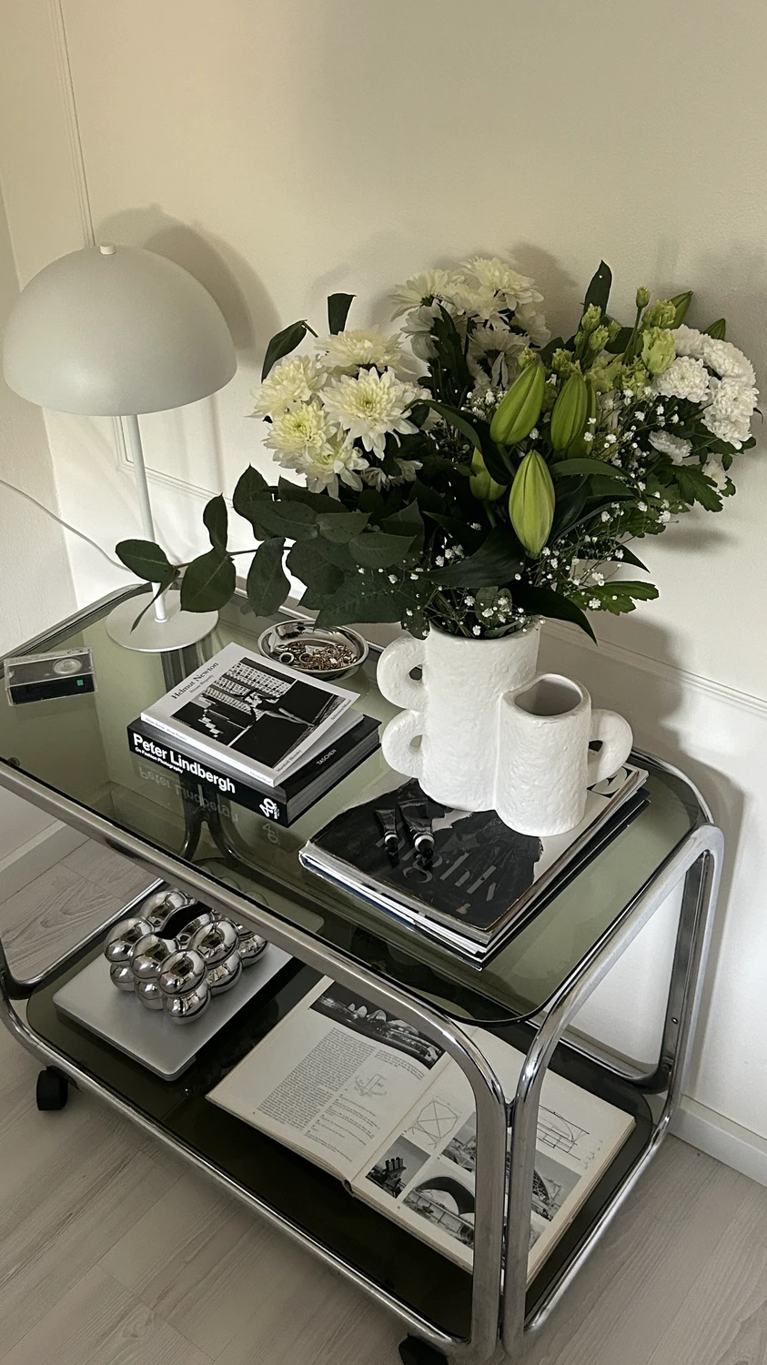 Spring decor ideas for living room showing a glass and chrome side table styled with coffee table books with fresh flowers arranged in a sculptural white vase, paired with a small table lamp, stacked magazines, and minimal decor accents that create a light, elegant seasonal vignette.