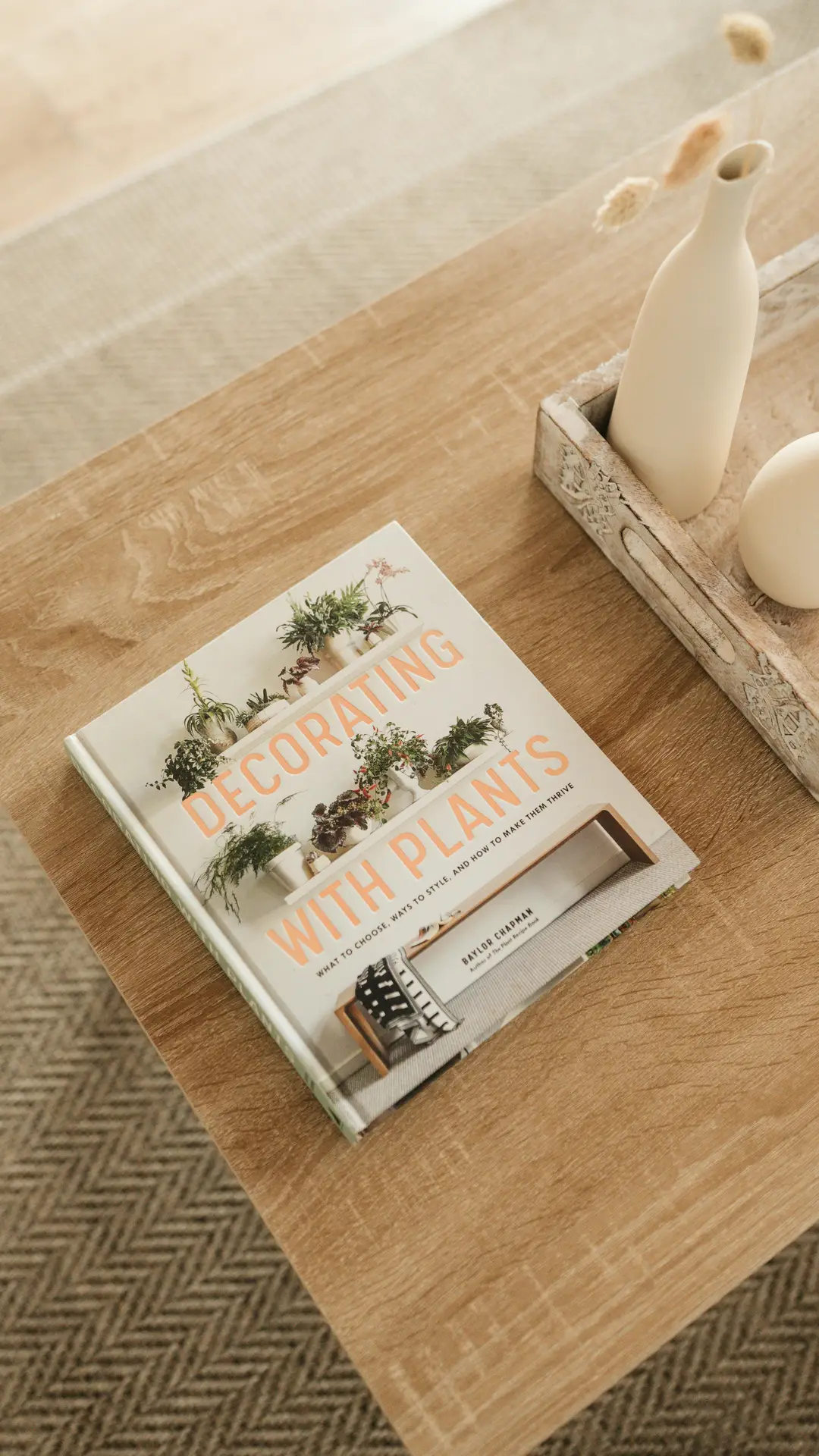 Spring decor ideas for living room featuring coffee table books arranged on a warm wooden surface, with a plant-themed hardcover placed neatly beside a rustic tray holding minimalist ceramic vases, creating a calm, natural styling with soft textures and light tones.