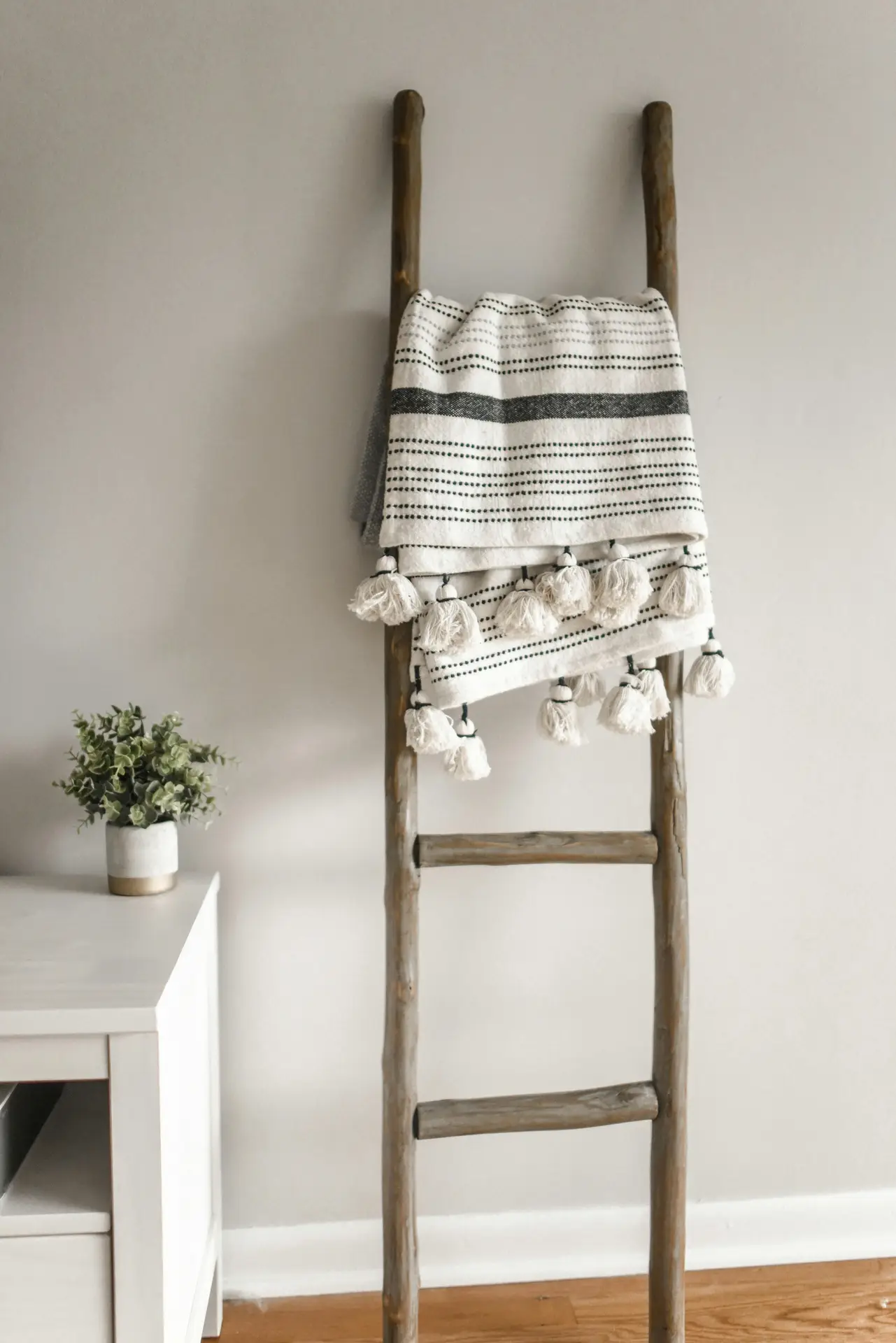 Neutral spring decor ideas for living room featuring a soft cotton blanket with subtle stripes and tassels draped over a rustic wooden ladder, placed against a light wall with a small side table and potted greenery nearby, creating a calm and airy seasonal styling.