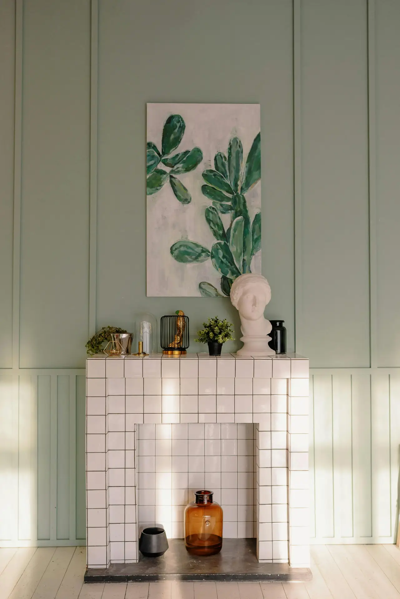 Spring decor ideas for living room showing how to decorate your fireplace for spring with a tiled mantel styled using small decorative objects, greenery, glass vases, and a sculptural bust, finished with botanical wall art against soft green paneling for a fresh, seasonal atmosphere.
