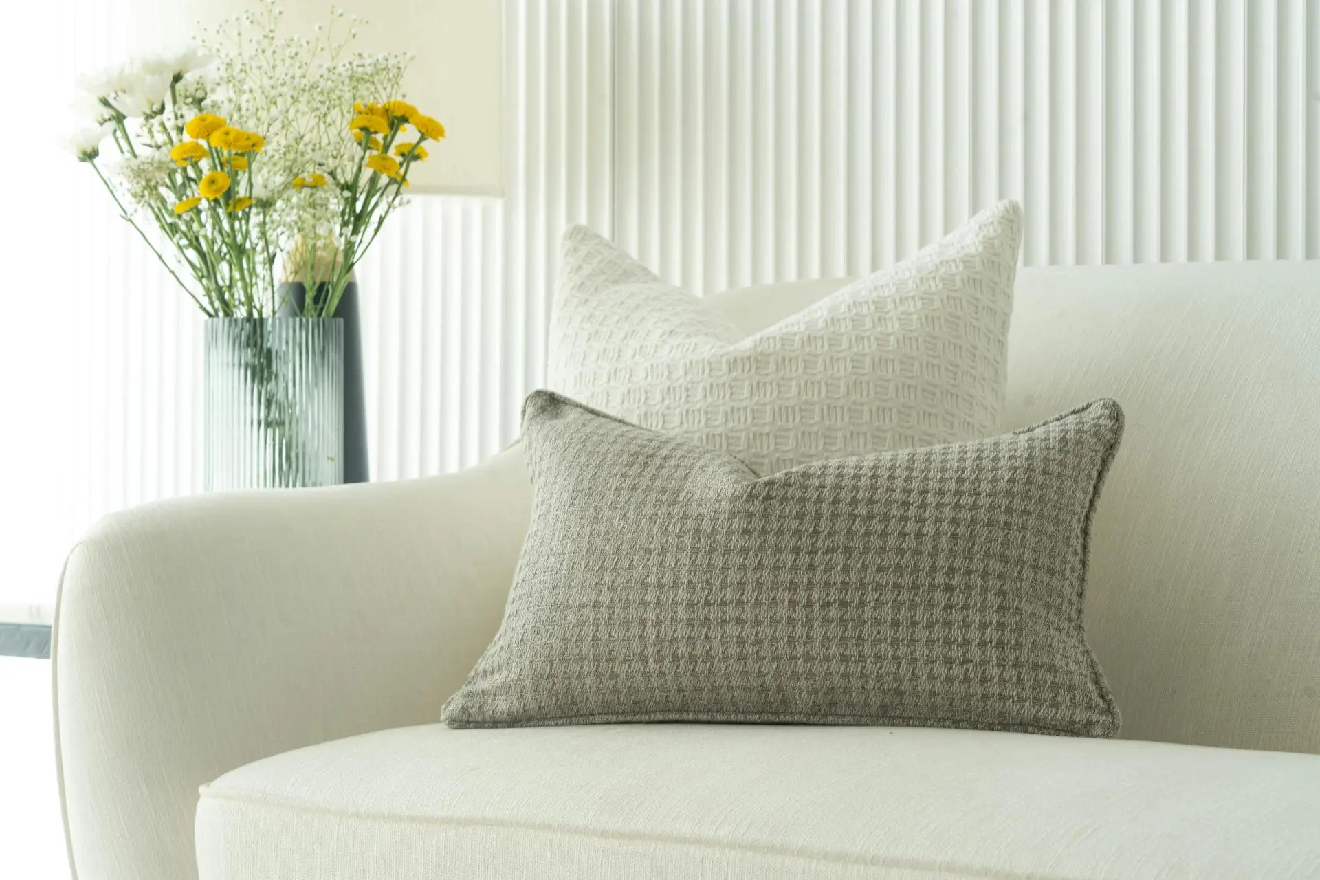 Elegant spring decor ideas for living room featuring neutral cushions layered on a light upholstered sofa, paired with a subtle textured wall and a glass vase filled with fresh yellow and white flowers in the background.