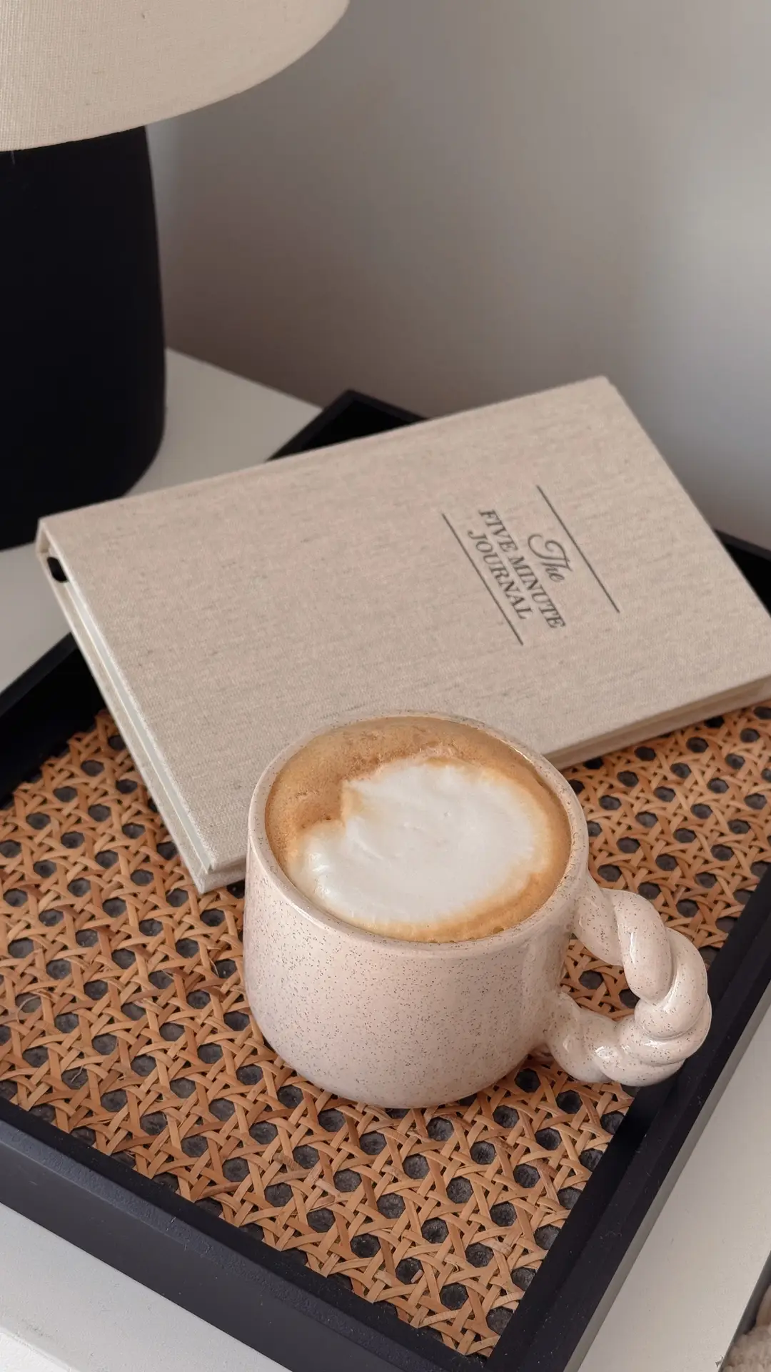 Cozy spring decor ideas for living room featuring a rattan tray styled with a neutral ceramic coffee mug, a soft linen-covered journal, and a table lamp, creating a calm, layered look with natural textures and a relaxed everyday feel.