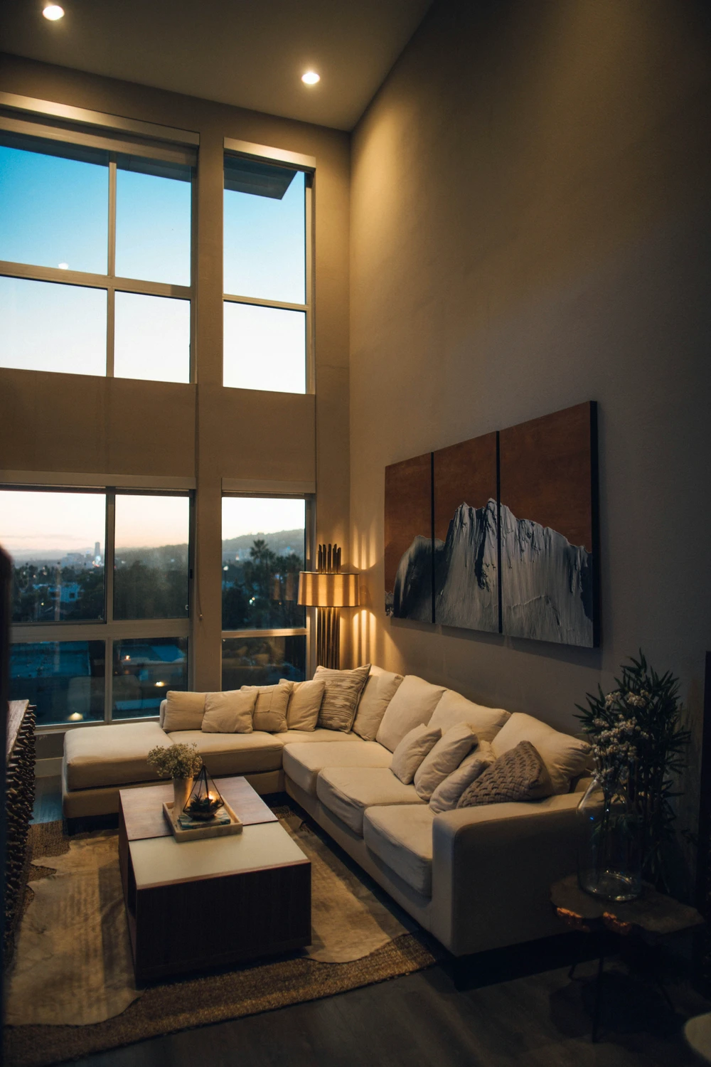 Modern double-height living room at dusk with floor-to-ceiling windows, a large cream sectional sofa, and a wooden coffee table styled with candles and flowers. Above the couch, a striking multi panel art work featuring a mountain landscape adds depth and texture, offering inspiring living room decor ideas for walls in a warm, softly lit contemporary space.
