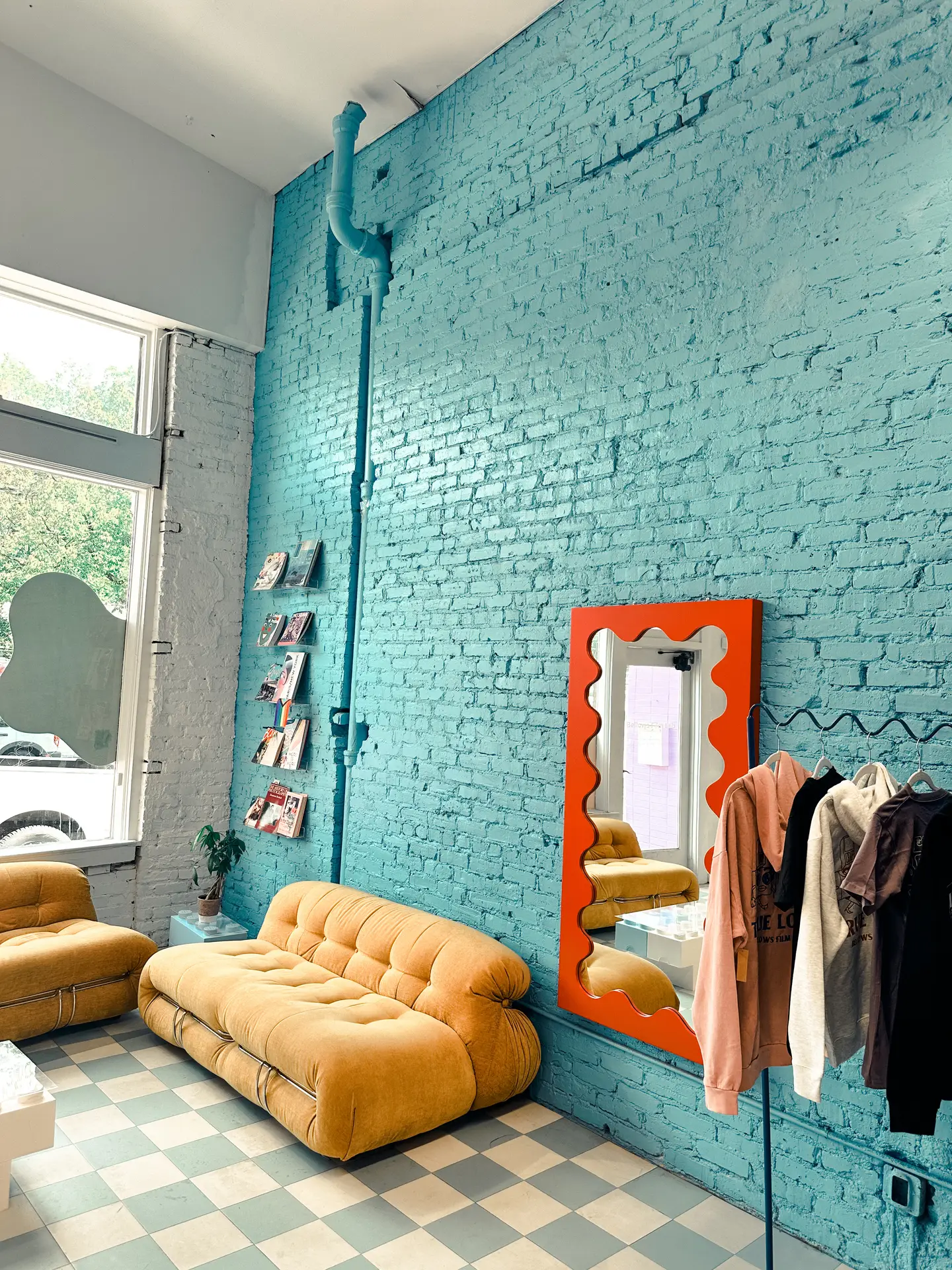An airy interior featuring living room paint color ideas with an aqua blue exposed brick wall, complemented by a mustard-yellow sofa, a sculptural mirror with a bold frame, a clothing rack, and soft daylight coming through large windows over a patterned floor.