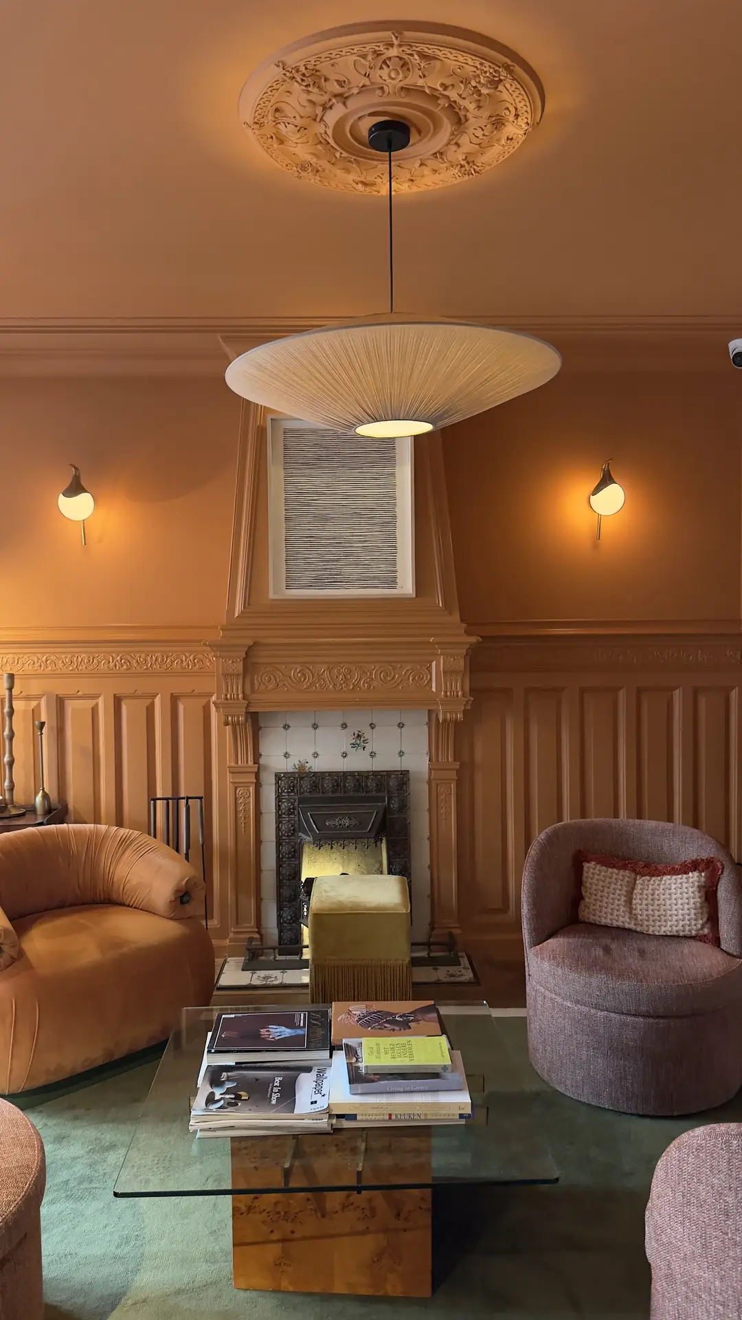 A cozy interior showcasing living room paint color ideas in a rich burnt orange tone, featuring ornate wall paneling, a decorative fireplace, soft upholstered armchairs in warm neutrals, ambient wall sconces, and a sculptural pendant light that adds depth and character to the space.