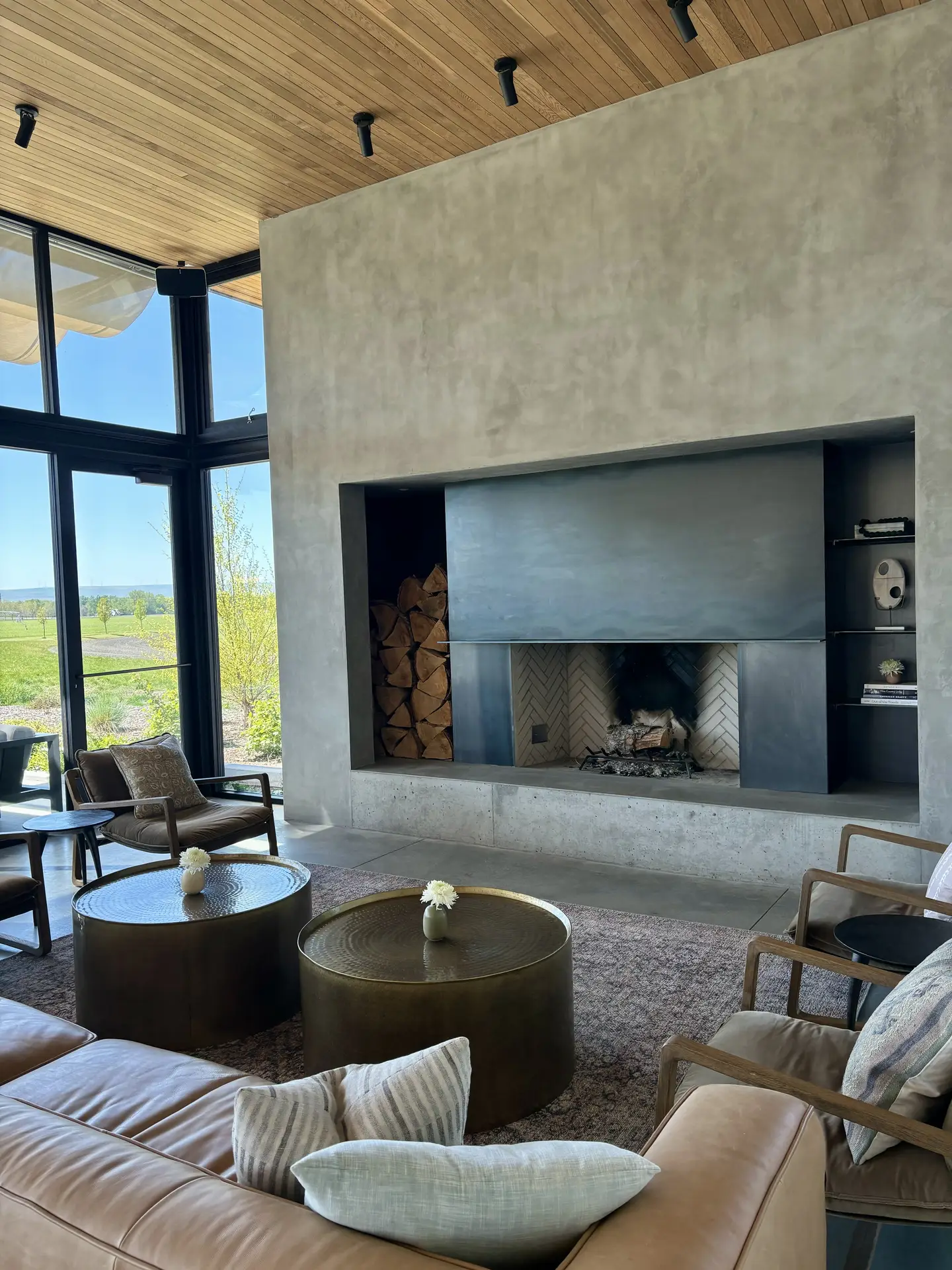 An airy interior showcasing living room paint color ideas with cement wash grey walls, featuring a modern fireplace set into textured concrete, warm leather seating, round metal coffee tables, and large floor-to-ceiling windows that bring natural light into the relaxed, contemporary space.