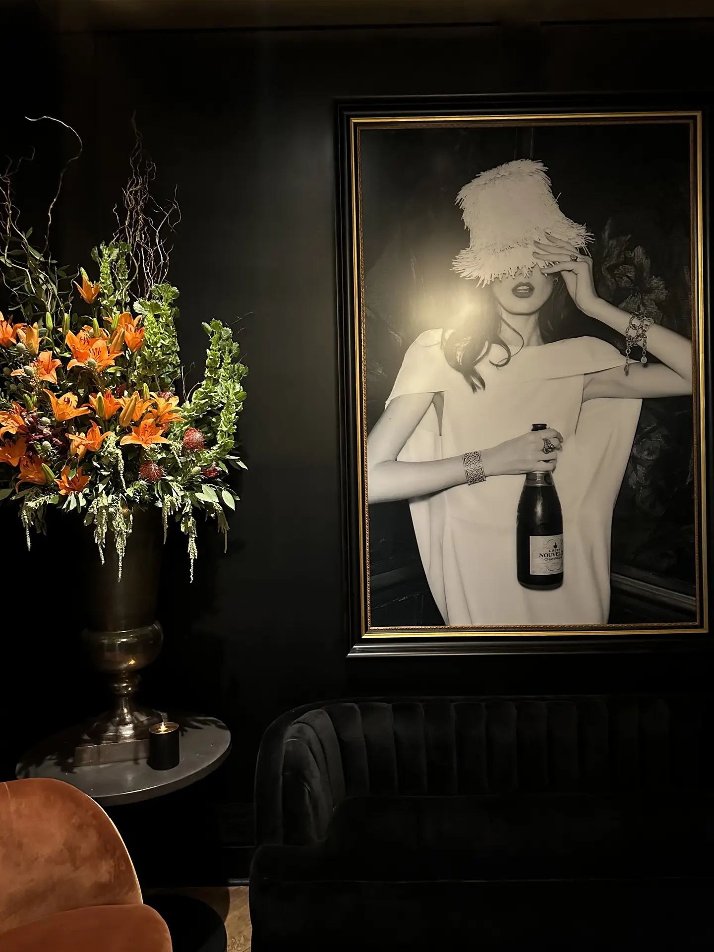 A dramatic interior scene presenting living room paint color ideas with deep black walls, featuring a moody setting with a dark velvet armchair, a round side table holding a lush floral arrangement, soft candlelight, and a large framed black-and-white portrait that adds contrast and elegance to the richly toned space.