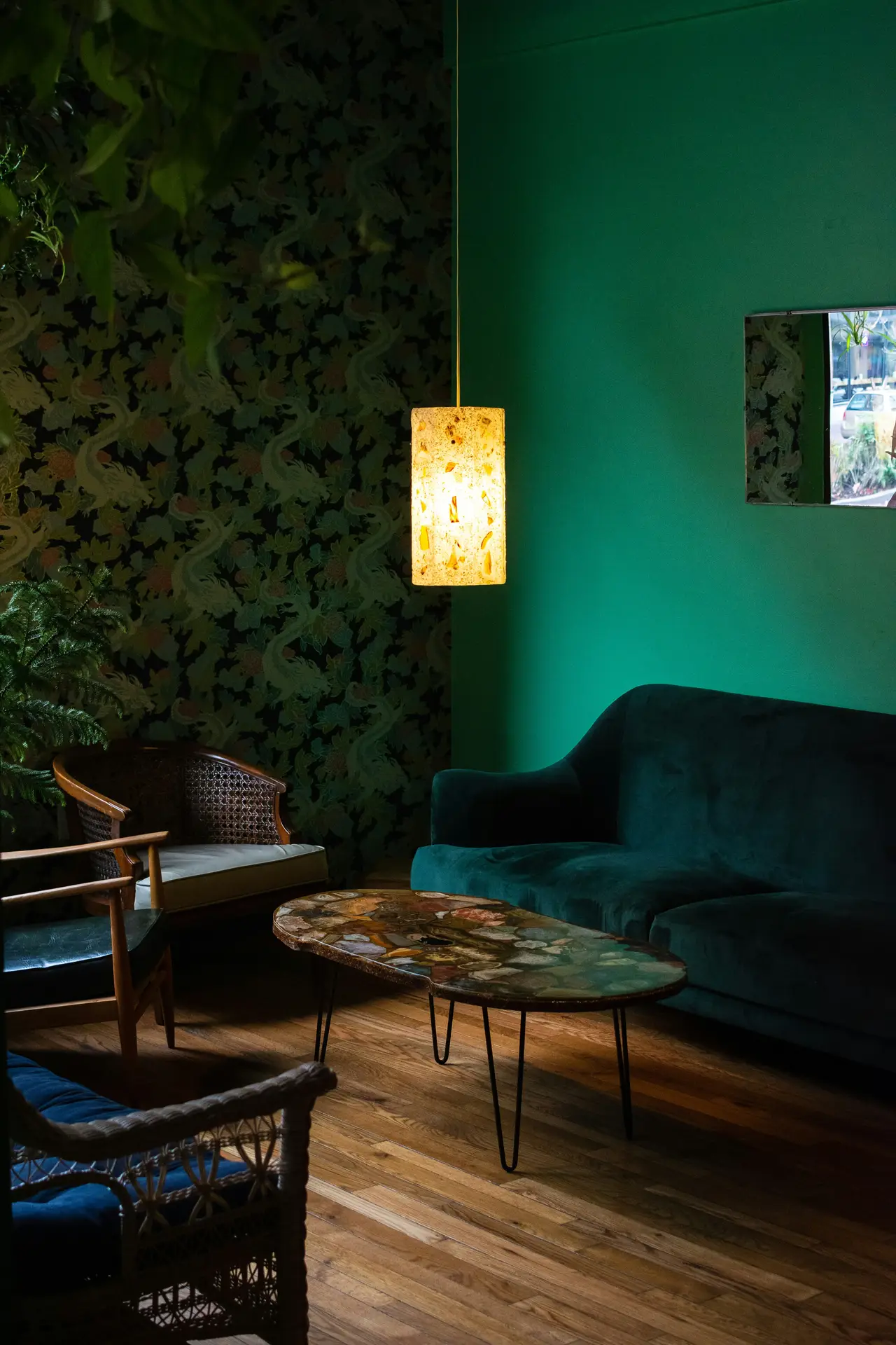 A moody interior scene highlighting living room paint color ideas with emerald green walls, featuring a deep green velvet sofa, patterned wallpaper accents, a sculptural pendant light casting warm glow, mid-century style chairs, a stone-topped coffee table, and rich wooden flooring that creates an intimate, sophisticated atmosphere.