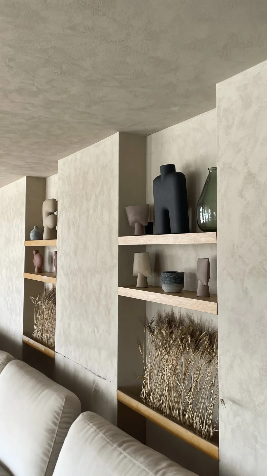 A calm, sculptural interior showcasing living room paint color ideas with a limewash beige finish, featuring textured walls, built-in niches with light wood shelves, neutral ceramic vases, and dried grasses that add depth, softness, and an earthy, modern feel to the space.