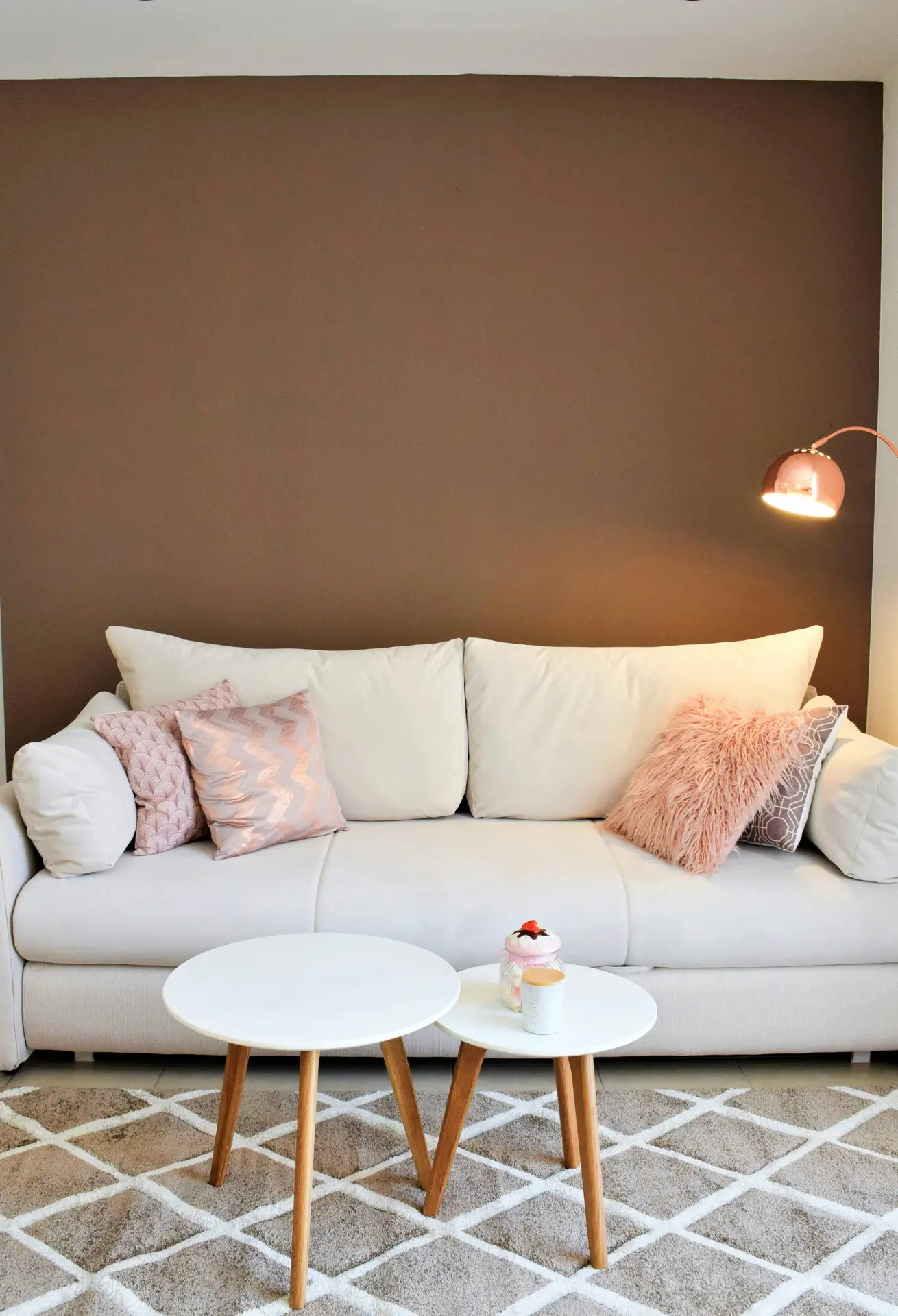 A cozy setup showcasing living room paint color ideas with a mocha brown accent wall behind a soft white sofa, styled with blush and textured throw pillows, paired with light wood nesting tables, a patterned rug, and a warm wall-mounted reading lamp for a calm, inviting feel.