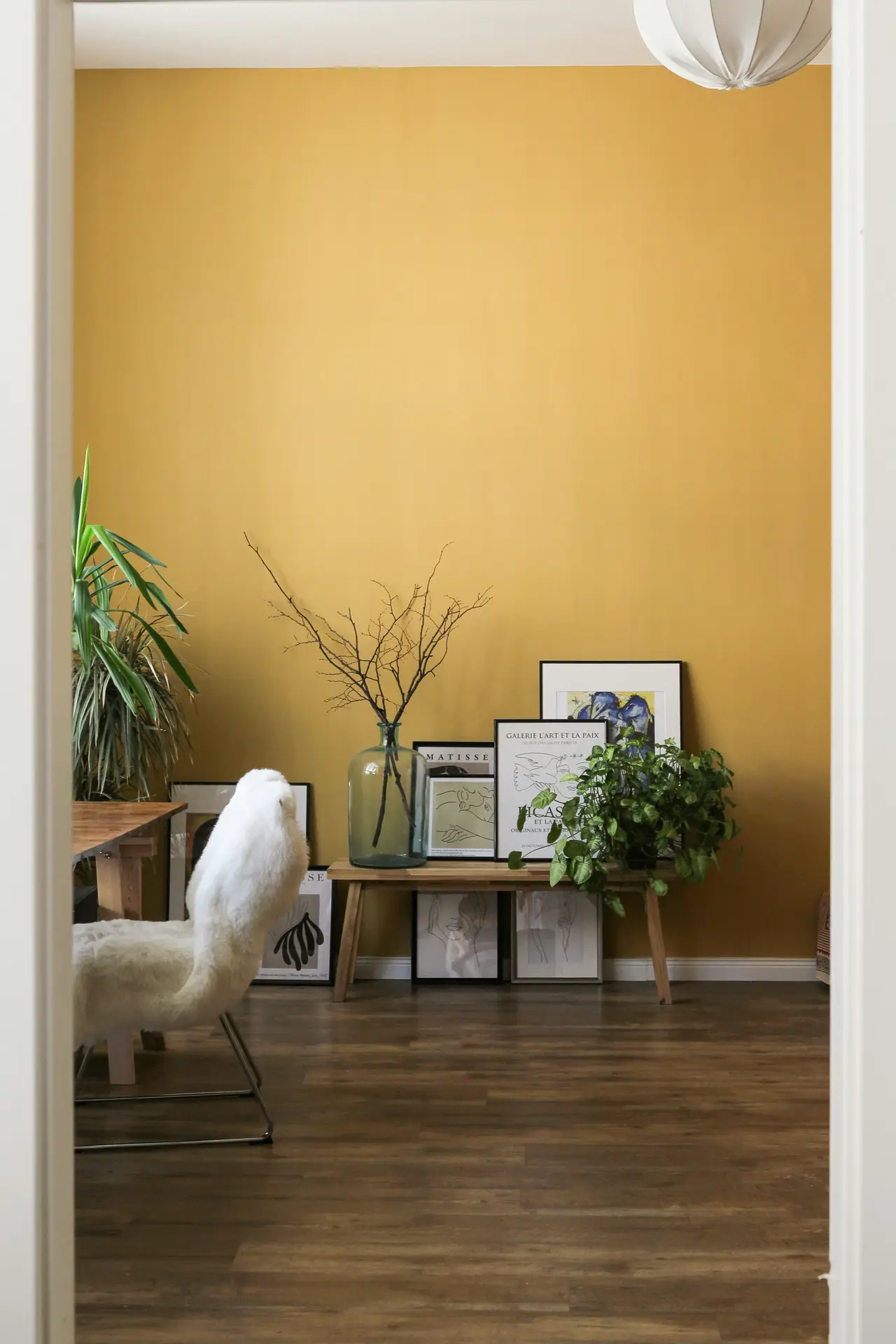 A cozy interior scene showcasing living room paint color ideas with a mustard yellow wall, featuring a simple wooden bench styled with framed art, a glass vase with branches, lush green plants, and a soft accent chair that adds warmth and character to the space.