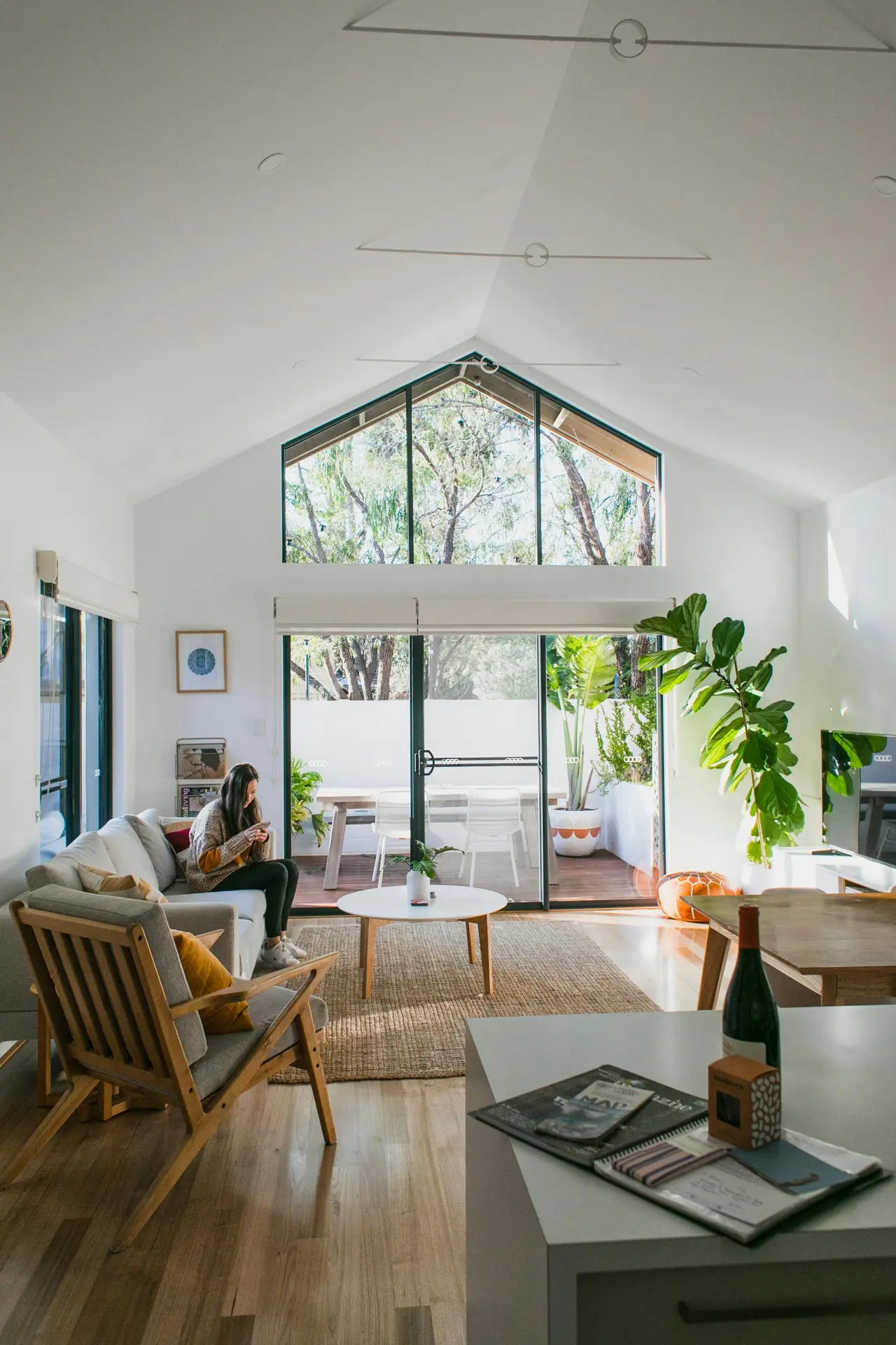 An open, light-filled interior showcasing living room paint color ideas with white walls, featuring a vaulted ceiling, large glass doors opening to a terrace, natural wood furniture, a cozy seating area with neutral textiles, indoor plants, and soft daylight creating a relaxed, modern atmosphere.