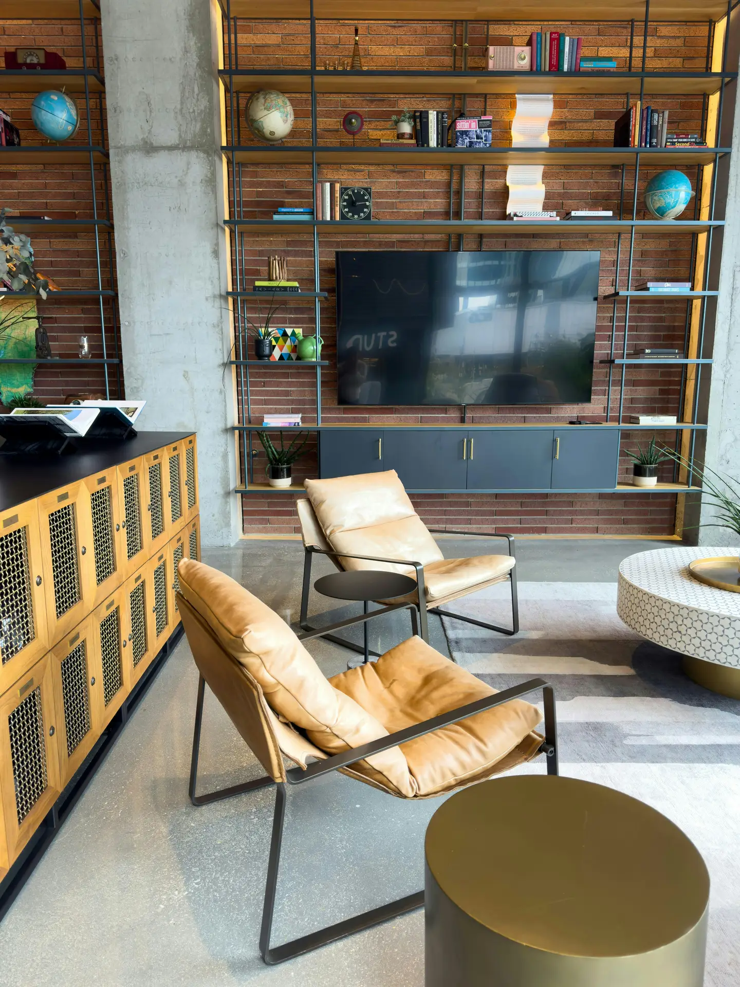Industrial-style living room featuring media wall ideas with a built-in media wall with open shelving set against an exposed brick backdrop. A flat-screen TV is centered within black metal shelves filled with books, globes, plants, and decorative objects, while a sleek gray cabinet runs below. Tan leather lounge chairs, round side tables, and a patterned coffee table complete the modern loft-inspired space with concrete floors and warm wood accents.