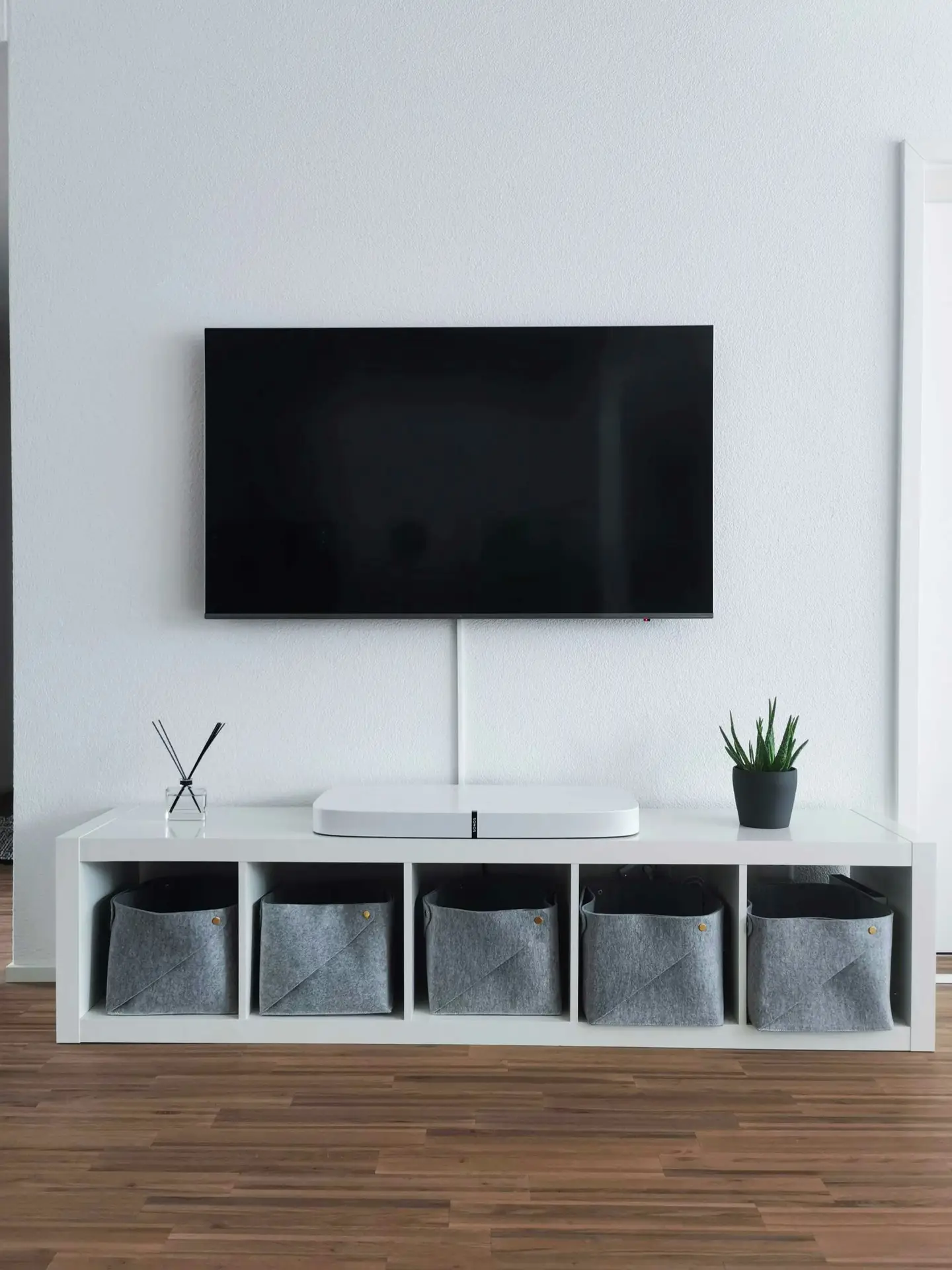 Minimalist living room setup featuring clean media wall ideas with a wall-mounted flat screen above a sleek white low board with boxes neatly arranged in open cubbies. The unit holds gray fabric storage bins, a small potted plant, and a diffuser, creating an organized and modern entertainment area against a light textured wall and warm wooden flooring.