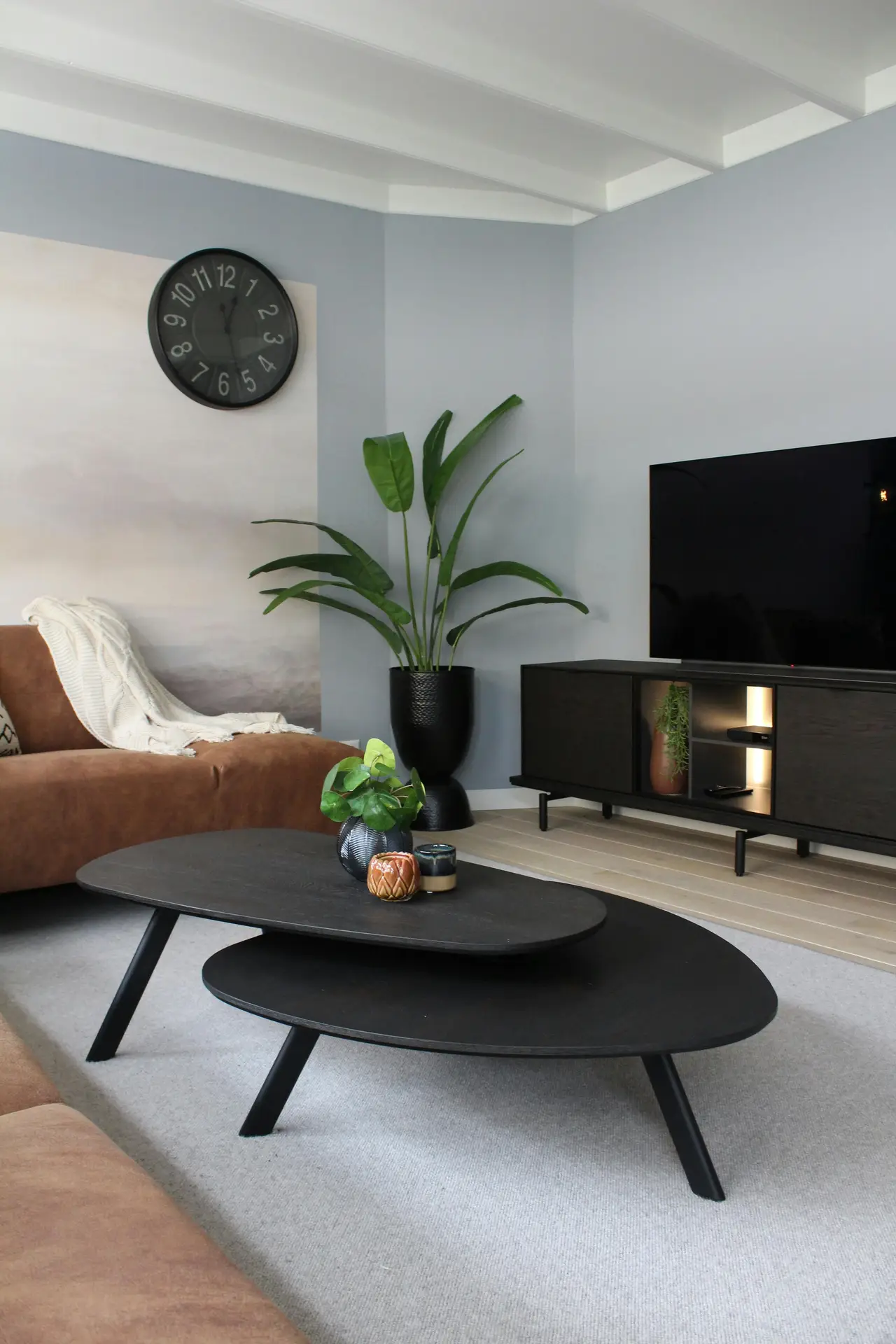 Cozy living room showcasing practical media wall ideas with a sleek low-profile media console in matte black beneath a flat-screen TV. The setup is styled with a tall leafy plant in a dark planter, layered black oval coffee tables, and warm brown sofas accented by soft throws. A large wall clock and muted blue-gray walls create a calm, contemporary atmosphere with subtle decorative touches.