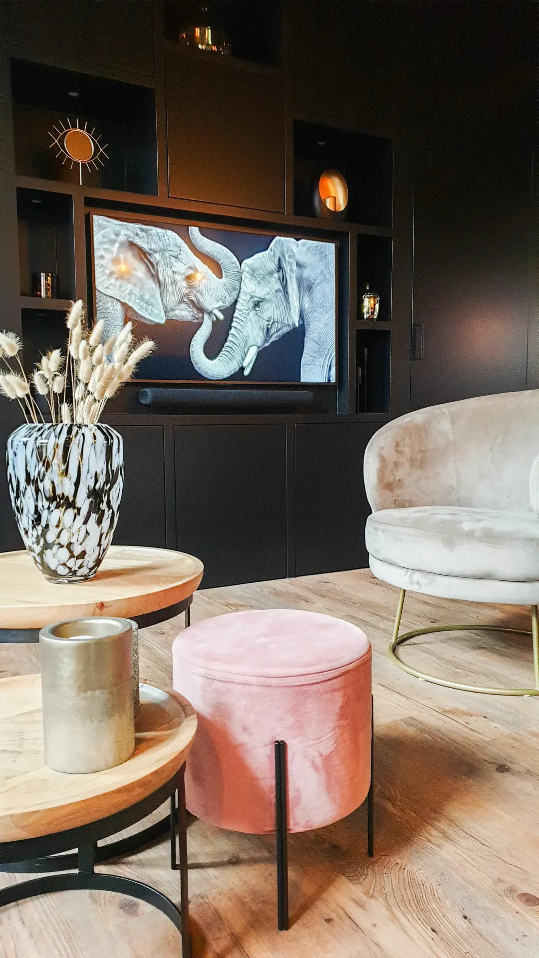Stylish living room featuring inspiring media wall ideas with a matte black built-in tv wall that frames the screen with custom shelving and subtle accent lighting. The television displays artwork of two elephants, set within dark cabinetry niches holding decorative objects. In the foreground, layered round wood tables, a pink velvet ottoman, a patterned vase with dried stems, and a curved upholstered chair create a cozy yet modern atmosphere.