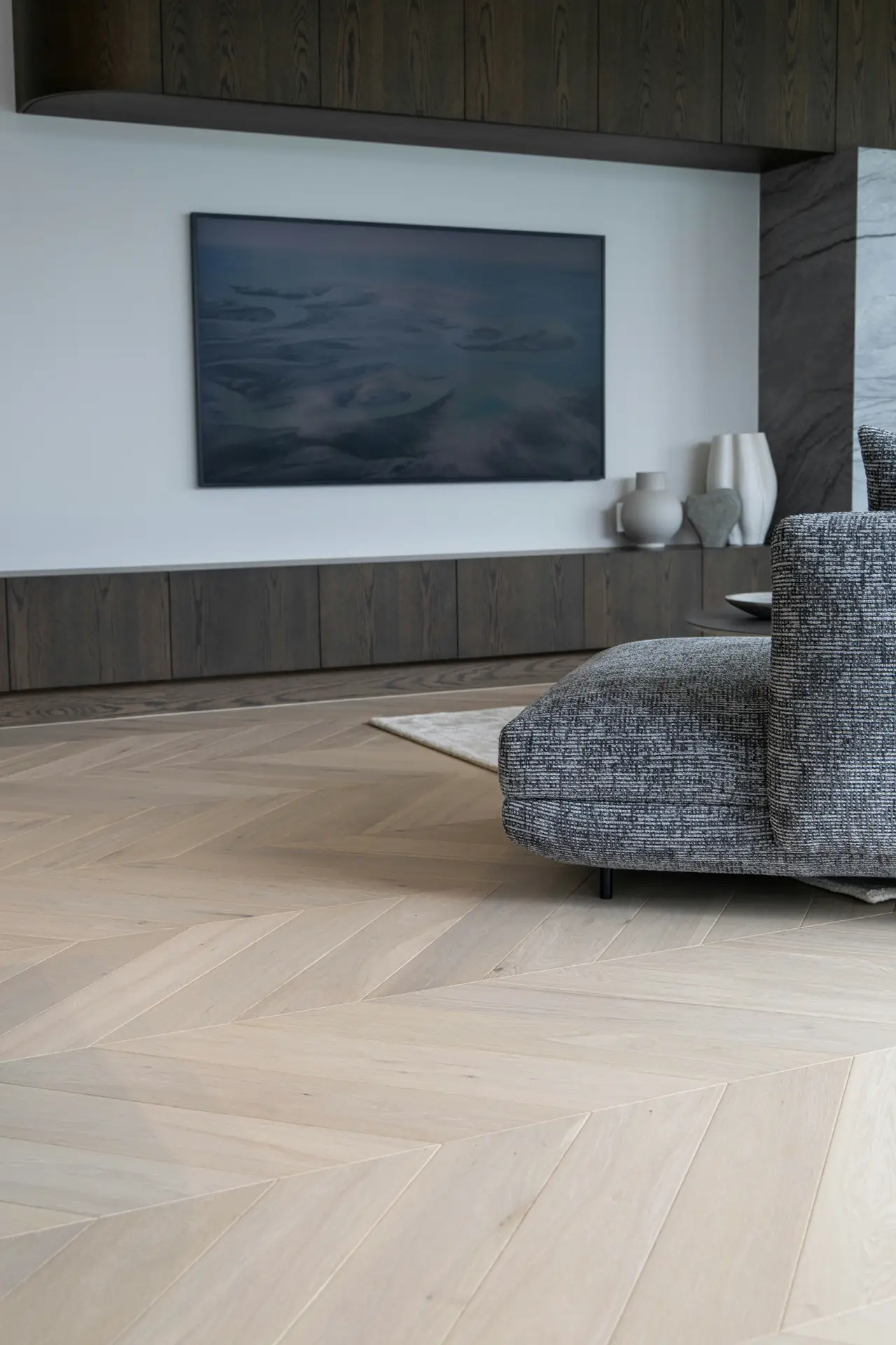 Contemporary living room featuring media wall ideas in a minimalist design, with a large wall-mounted screen set into a sleek wood-paneled unit and clean white backdrop. A low built-in cabinet runs beneath the display, styled with simple ceramic vases, while a textured gray lounge chair and light herringbone wood flooring complete the calm, modern atmosphere.