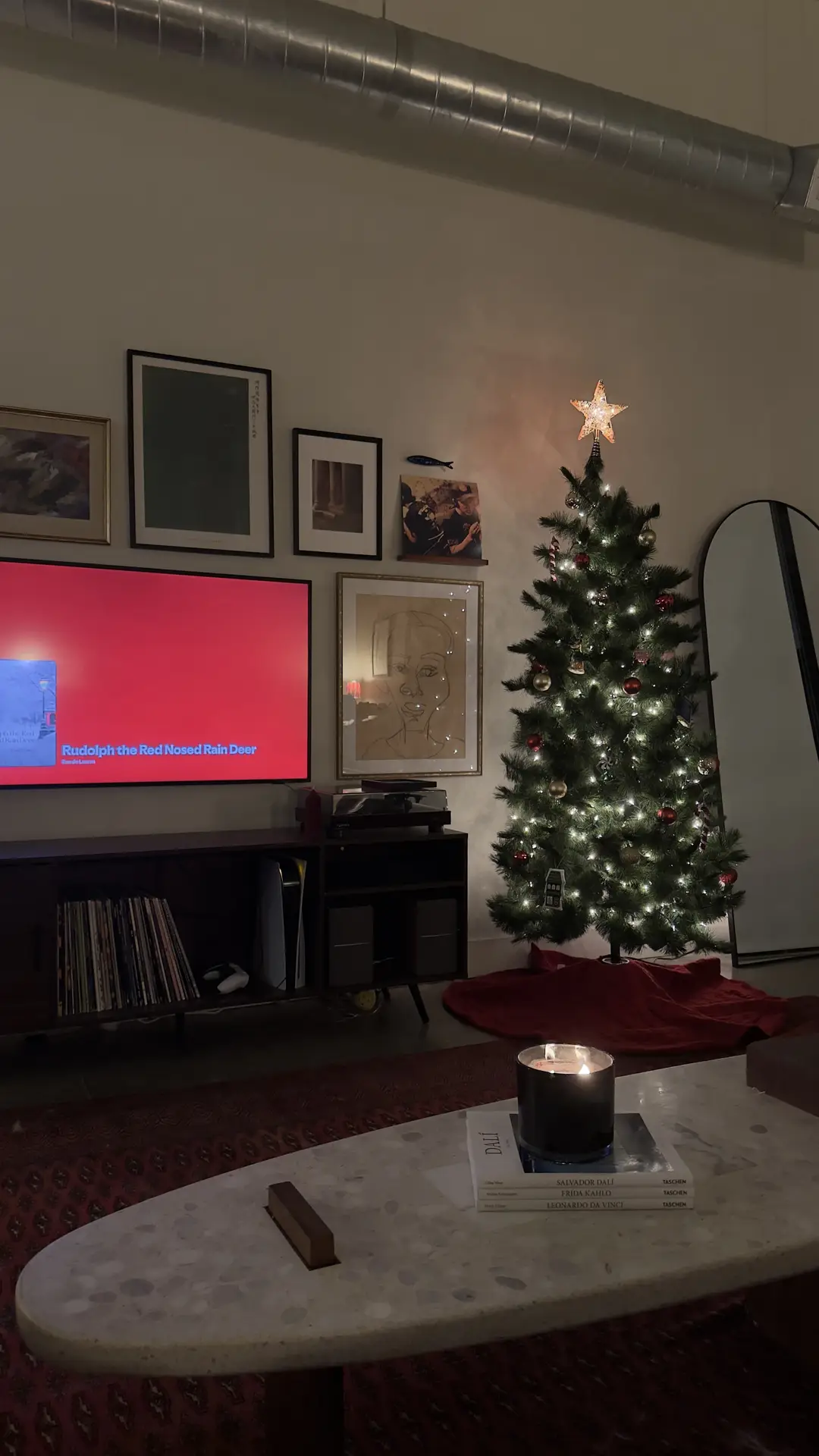Cozy living room at night featuring a decorated Christmas tree with warm string lights and a glowing star topper beside a sleek media console. The television is mounted on a stylish media wall with curated photo frames around tv, creating inspiring media wall ideas that blend art prints, framed photography, and ambient candlelight for a warm, personal atmosphere.