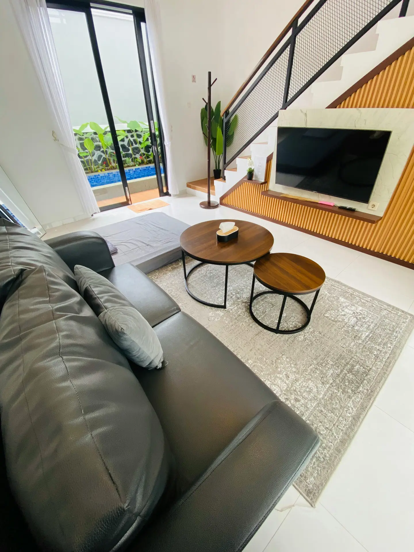 Contemporary living room showcasing creative media wall ideas with a staircase media wall design. A flat-screen TV is integrated into a marble and vertical wood slat feature beneath the stairs, paired with floating shelving. The space includes a black leather sofa, round nesting coffee tables on a textured rug, and sliding glass doors that open to a small outdoor pool area, bringing in natural light and greenery.