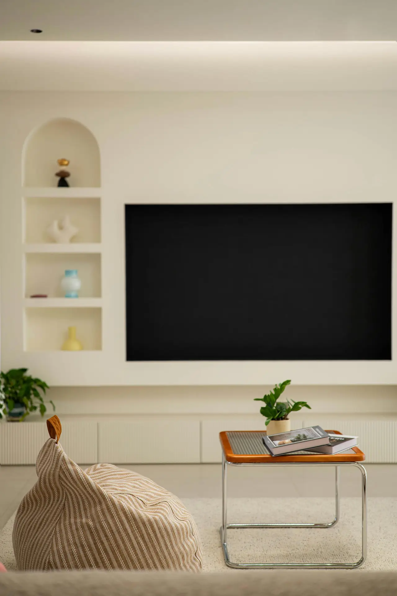 Contemporary living room showcasing creative media wall ideas with a wall-integrated tv with recessed shelving set into a smooth cream backdrop. The large flat-screen is framed by built-in niches featuring sculptural decor and colorful vases, while a long low cabinet runs beneath the display for streamlined storage. In the foreground, a striped floor cushion and a small chrome-and-wood side table with books and a potted plant add warmth to the clean, minimalist setting.