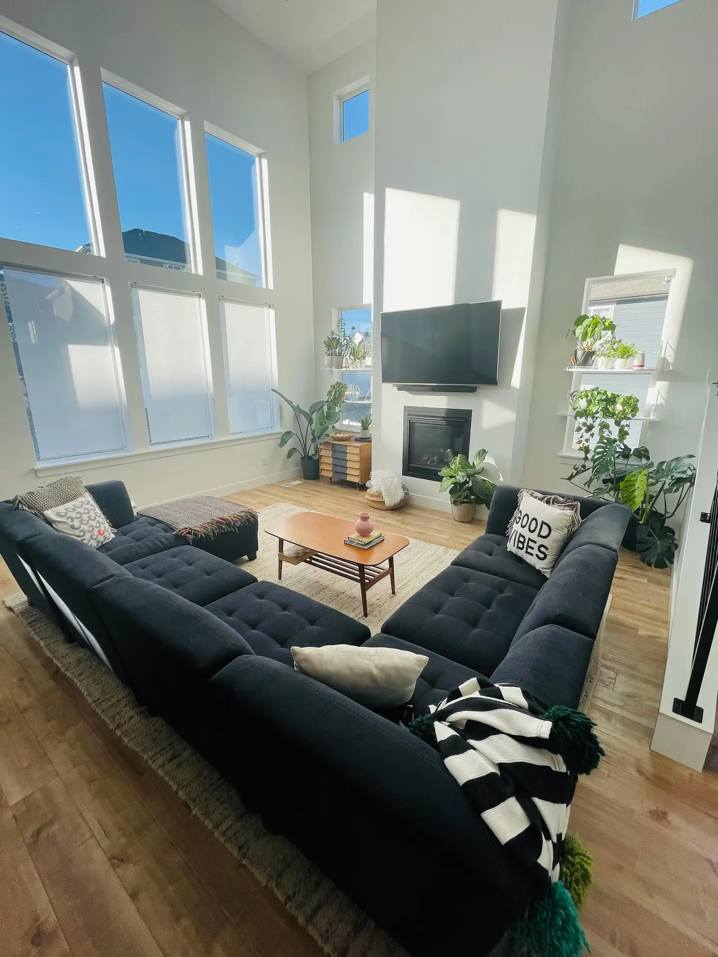 Spacious, light-filled living room featuring inspiring media wall ideas with a wall-mounted tv over cornerstone fireplace. Tall grid windows flood the space with sunlight, highlighting a fireplace beneath the screen. A large dark sectional sofa frames a mid-century style coffee table, while layered rugs and abundant green plants add warmth and texture to the airy, modern interior.