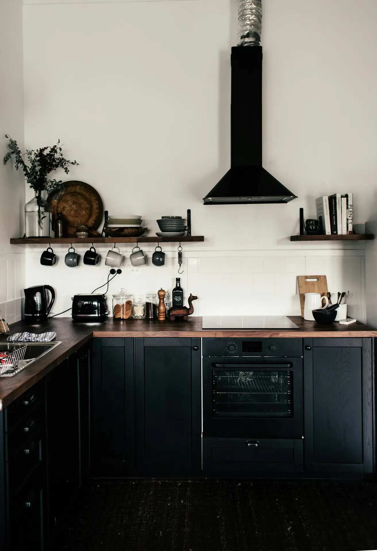 Moody yet functional corner of modern farmhouse kitchens featuring black cabinets paired with warm wooden countertops, open shelves holding dishes and mugs, a matte black range hood, and simple everyday appliances that balance modern contrast with cozy farmhouse character.
