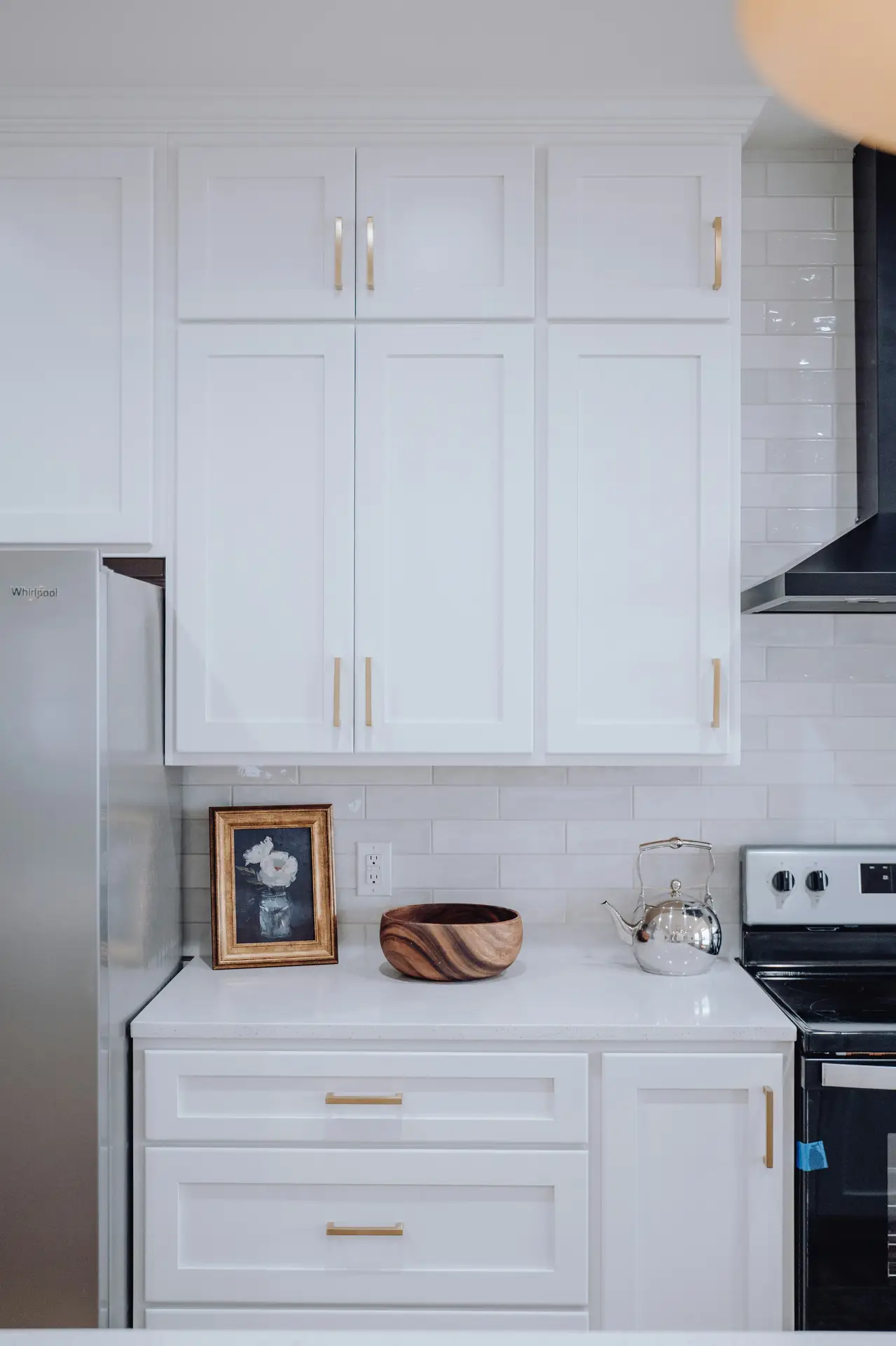 Clean and polished view of modern farmhouse kitchens, highlighting white shaker cabinets with brass handles, a glossy subway tile backsplash, and simple countertop decor like a wooden bowl and framed artwork, paired with stainless steel appliances for a timeless yet updated look.