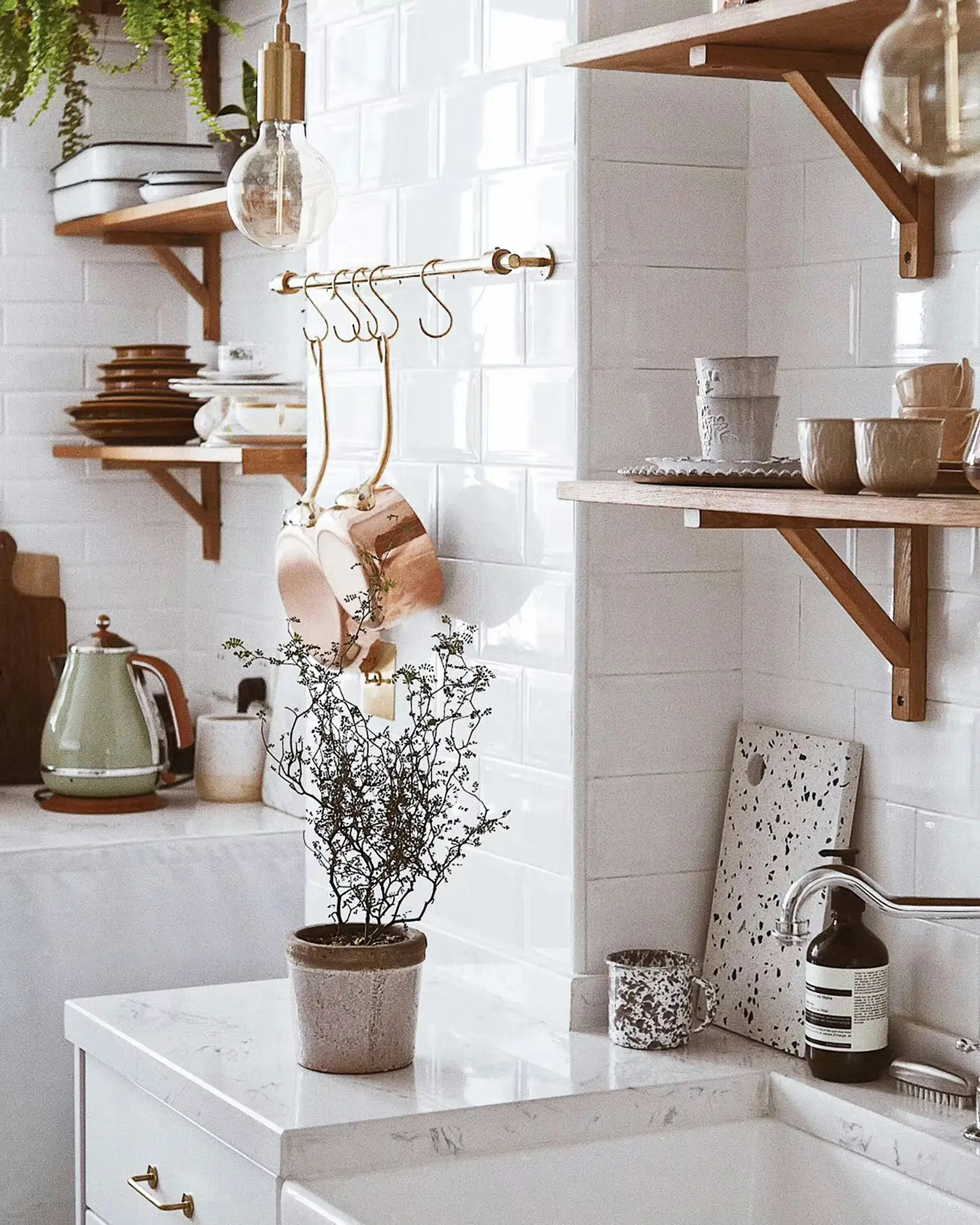 Light-filled detail of modern farmhouse kitchens showcasing ceramic tableware and vintage pots on open wooden shelves, with a white tile backsplash, brass hooks holding cookware, soft greenery, and simple countertop styling that blends practical storage with decorative charm.