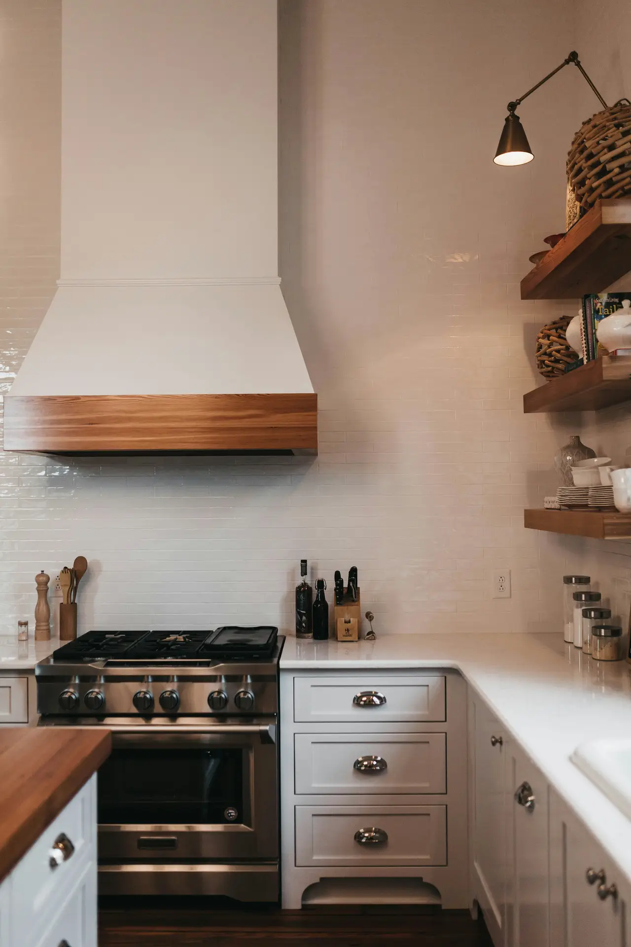 Warm and refined view of modern farmhouse kitchens combining wood and stone, featuring a white range hood with a wooden trim, open wooden shelves, a stone-style backsplash, and a stainless steel stove, all styled with simple kitchen essentials for a balanced, inviting atmosphere.