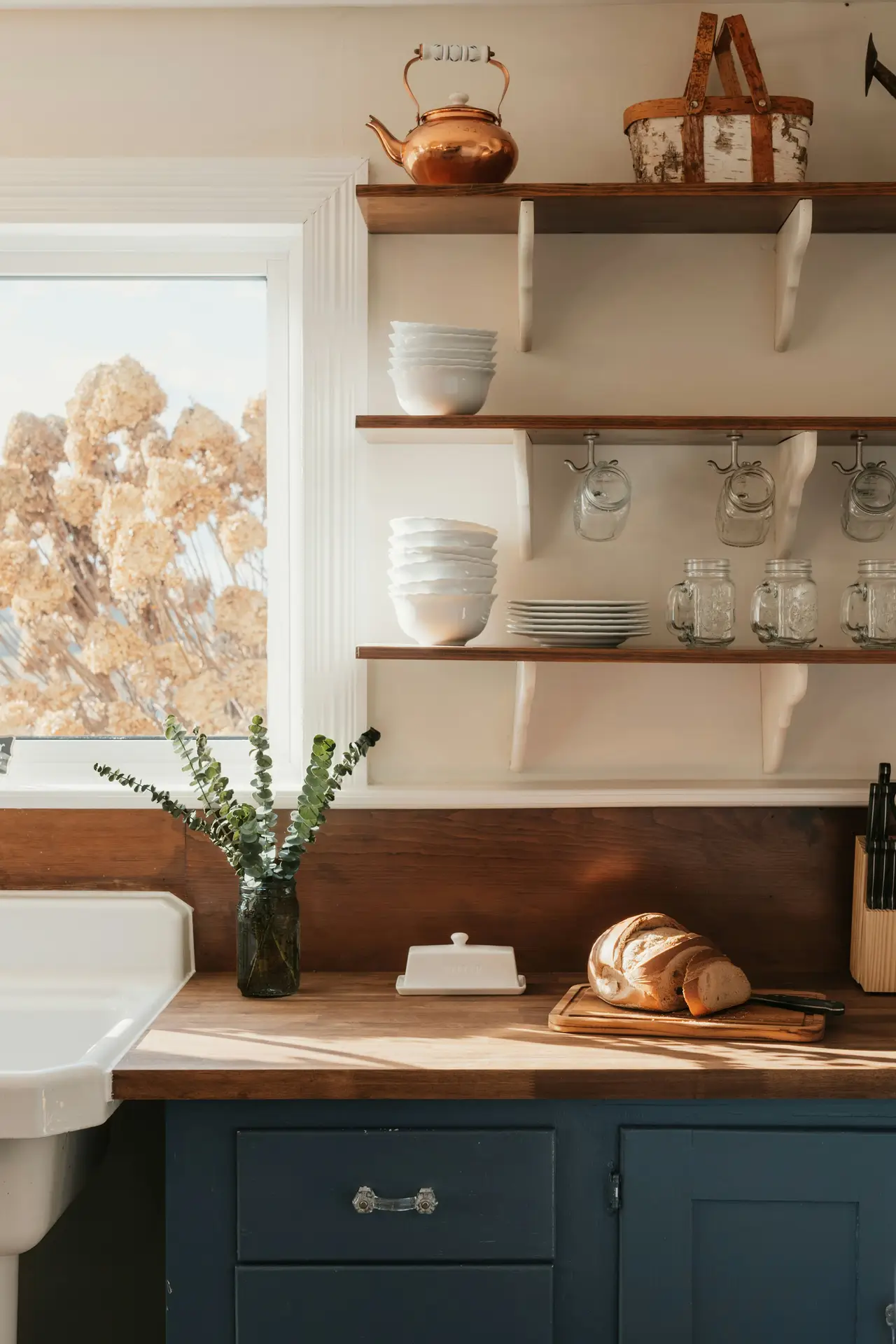 Cozy corner of modern farmhouse kitchens featuring dark blue cabinets paired with warm wooden countertops, open shelves filled with white dishes and glassware, a farmhouse sink, and rustic details like a copper kettle and fresh bread, all softly lit by natural daylight from the window.