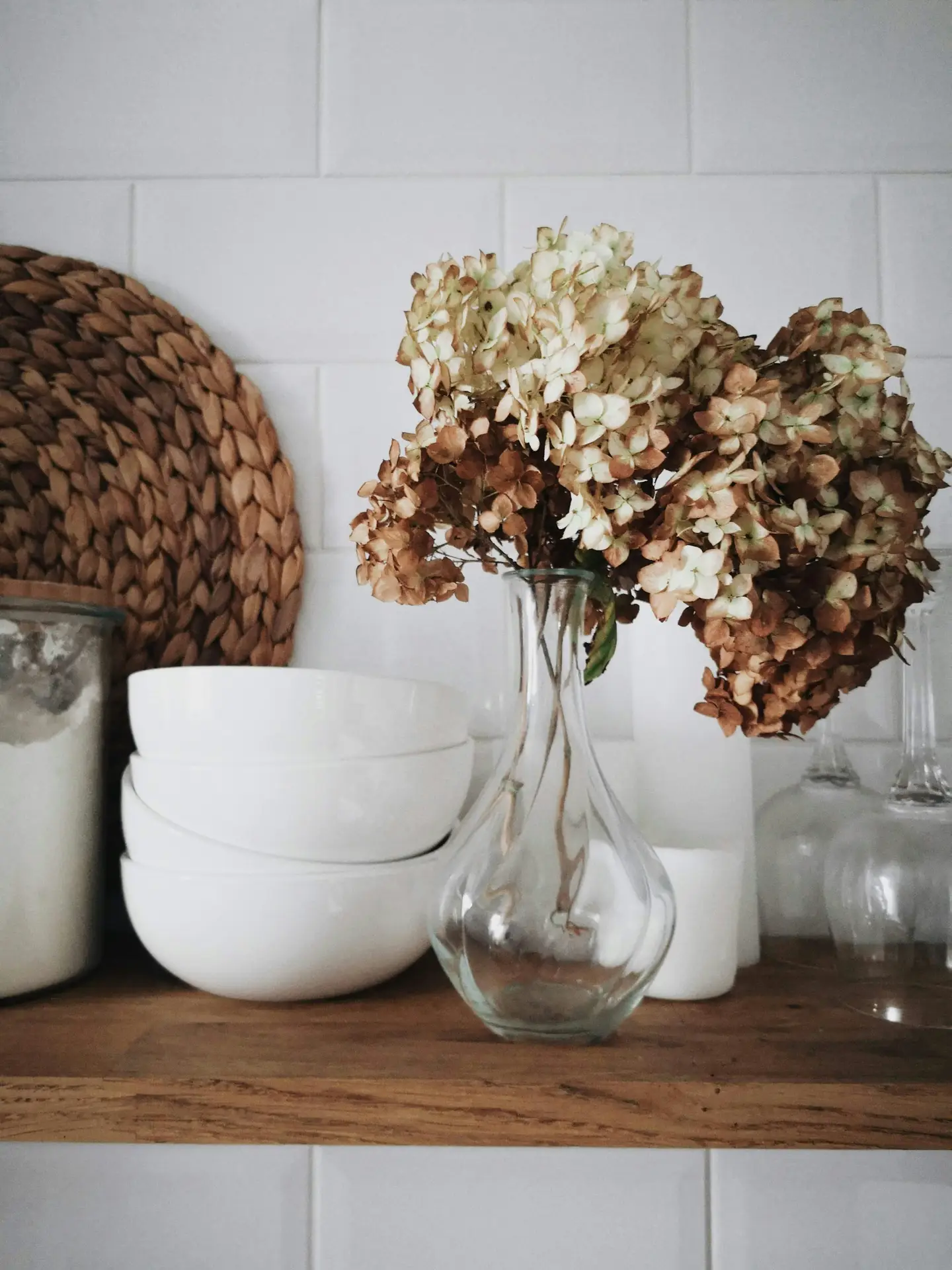 Simple shelf styling in modern farmhouse kitchens featuring dried flowers arranged in a clear glass vase, paired with stacked white bowls, woven accents, and glass containers against a classic white tile backdrop for a calm, natural look.