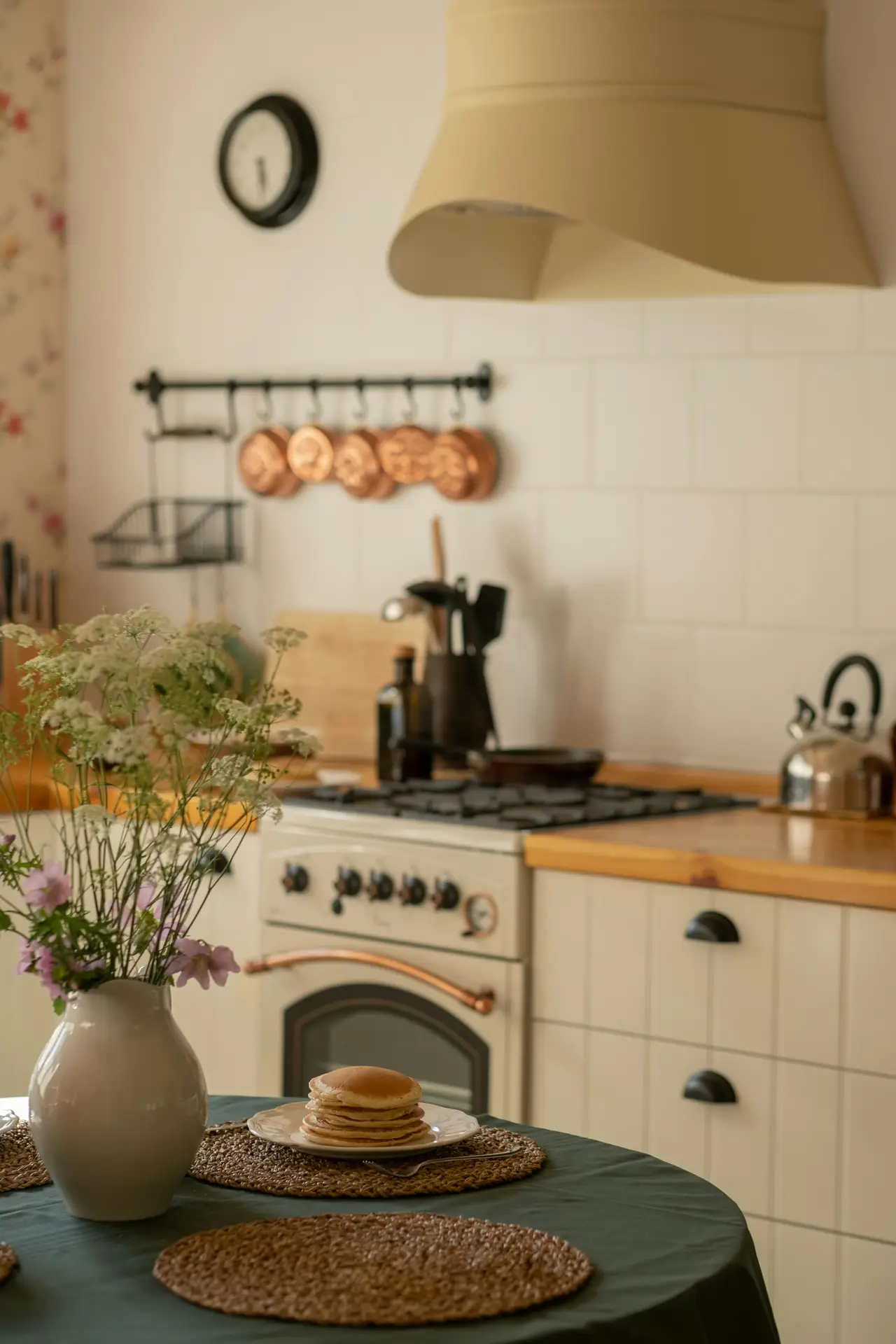 Soft and inviting scene of modern farmhouse kitchens, featuring fresh flowers in a simple vase on a dining table, warm wood countertops, cream cabinetry, copper cookware on the wall, and a classic stove setup that creates a cozy, lived-in kitchen atmosphere.