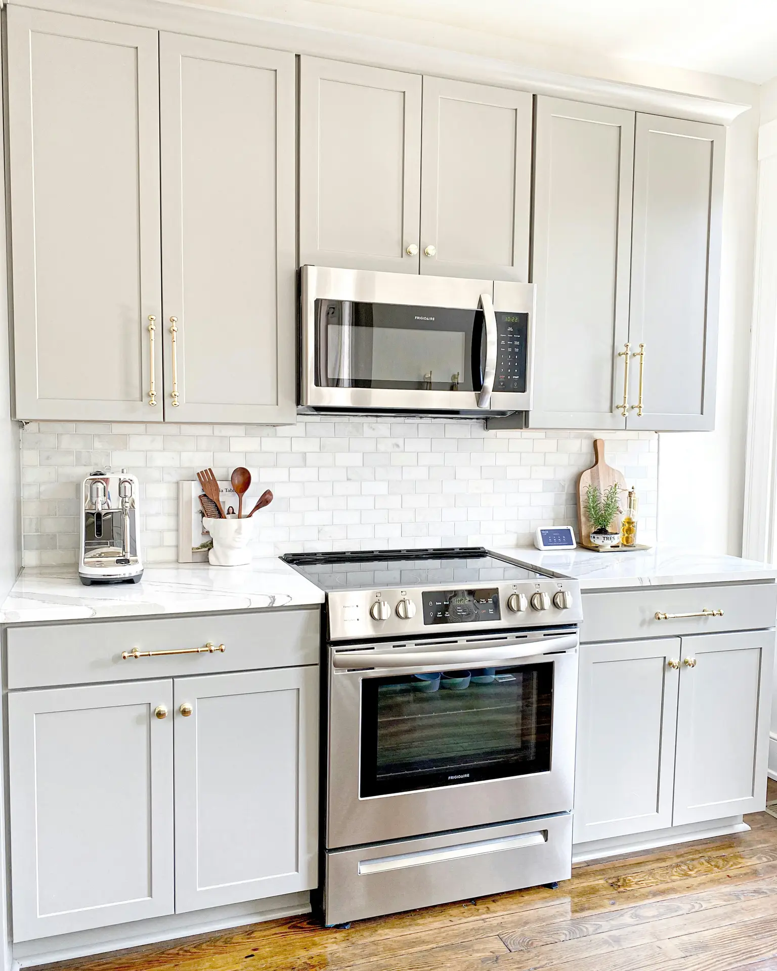 Bright and airy view of modern farmhouse kitchens featuring light grey cabinets with brass hardware, a white subway tile backsplash, marble-style countertops, and stainless steel appliances, styled with simple wooden accents and everyday kitchen tools for a clean yet welcoming look.