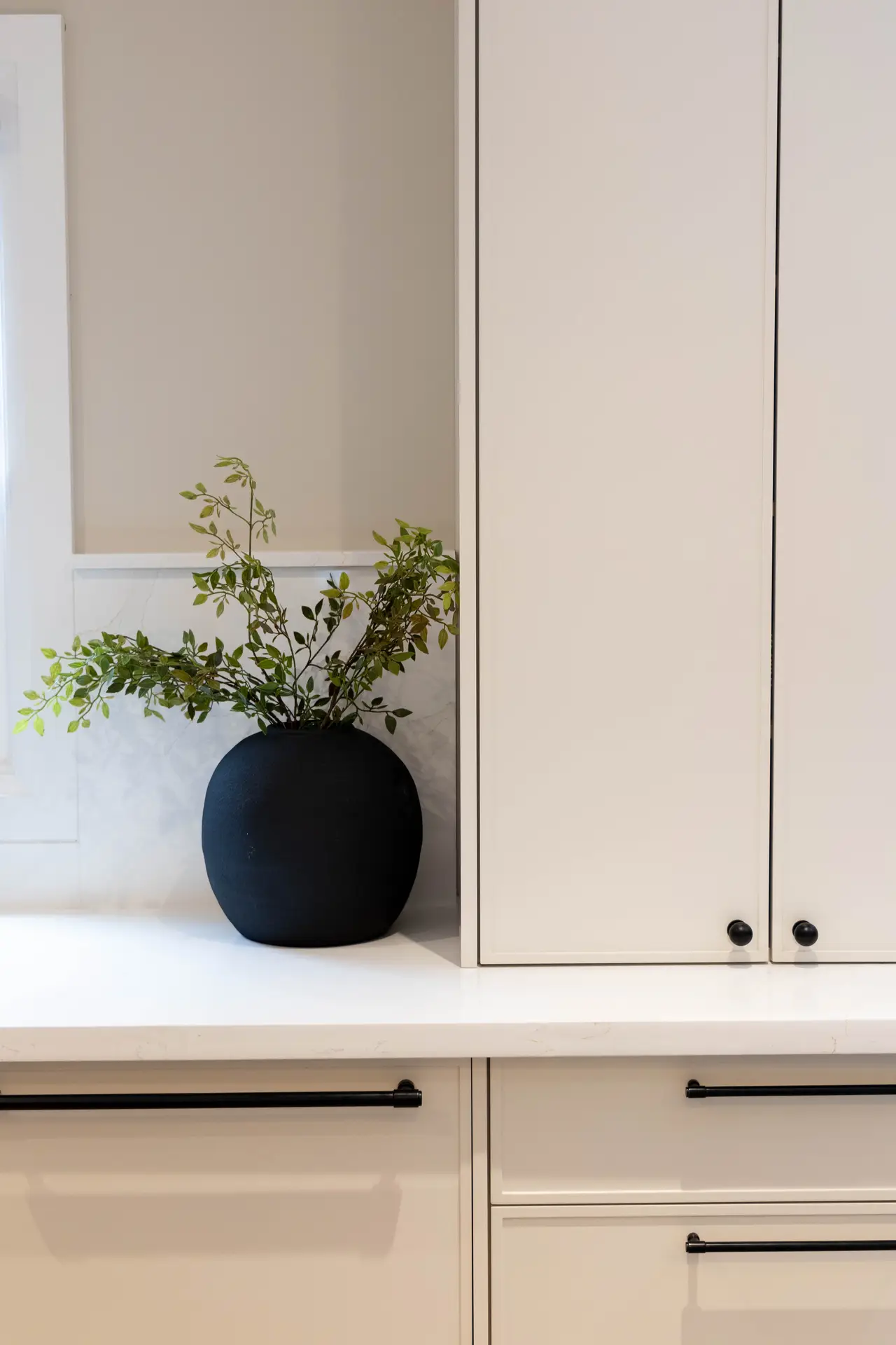 Clean and understated detail from modern farmhouse kitchens with a minimalist design, featuring smooth light-toned cabinetry, slim black hardware, a white countertop, and a simple black vase with greenery that adds a subtle natural accent without visual clutter.