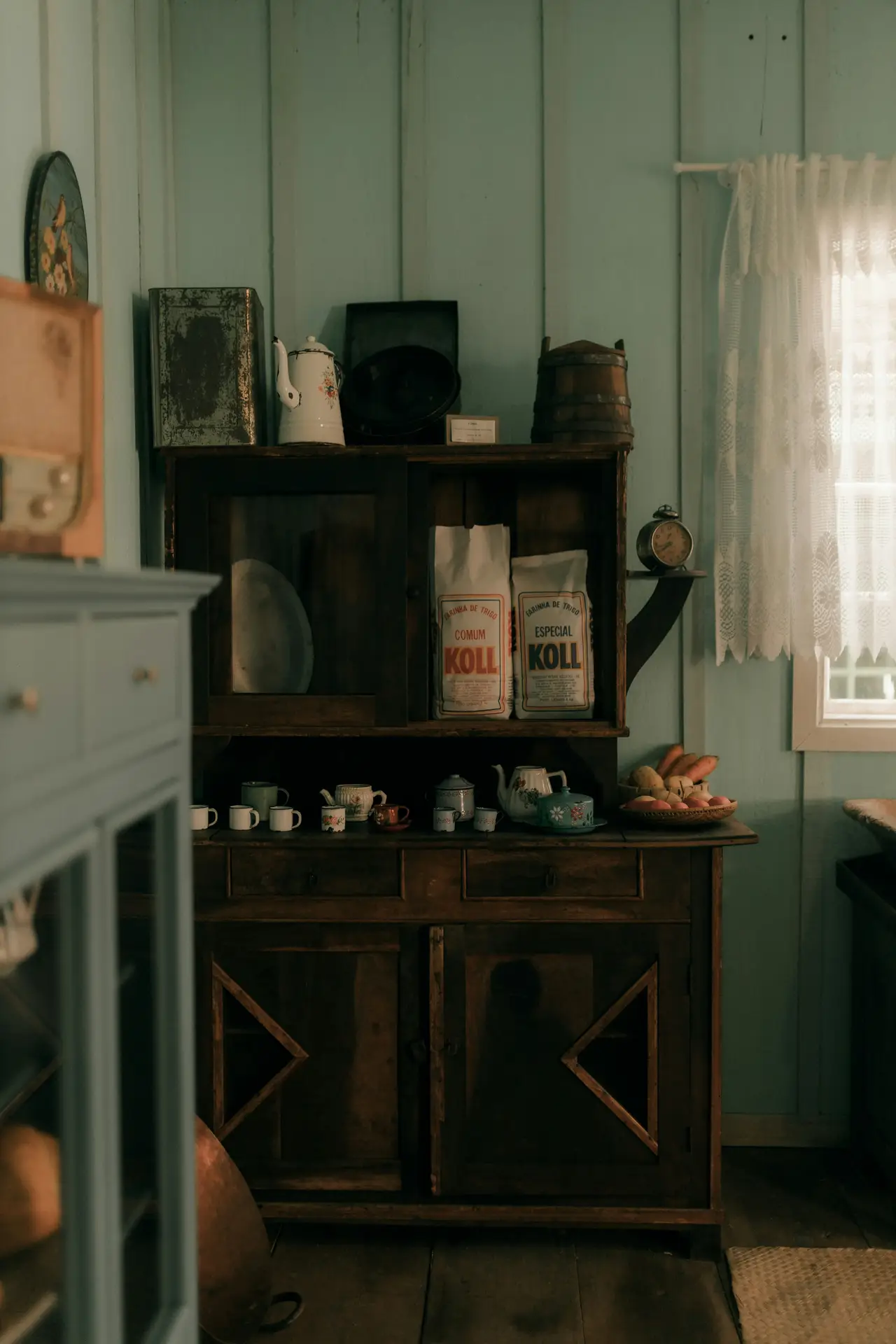 Moody and nostalgic corner of modern farmhouse kitchens, featuring a vintage cabinet filled with old kitchenware, flour bags, teacups, and rustic containers, set against painted wooden walls and soft daylight filtering through a lace curtain for a timeless, collected feel.