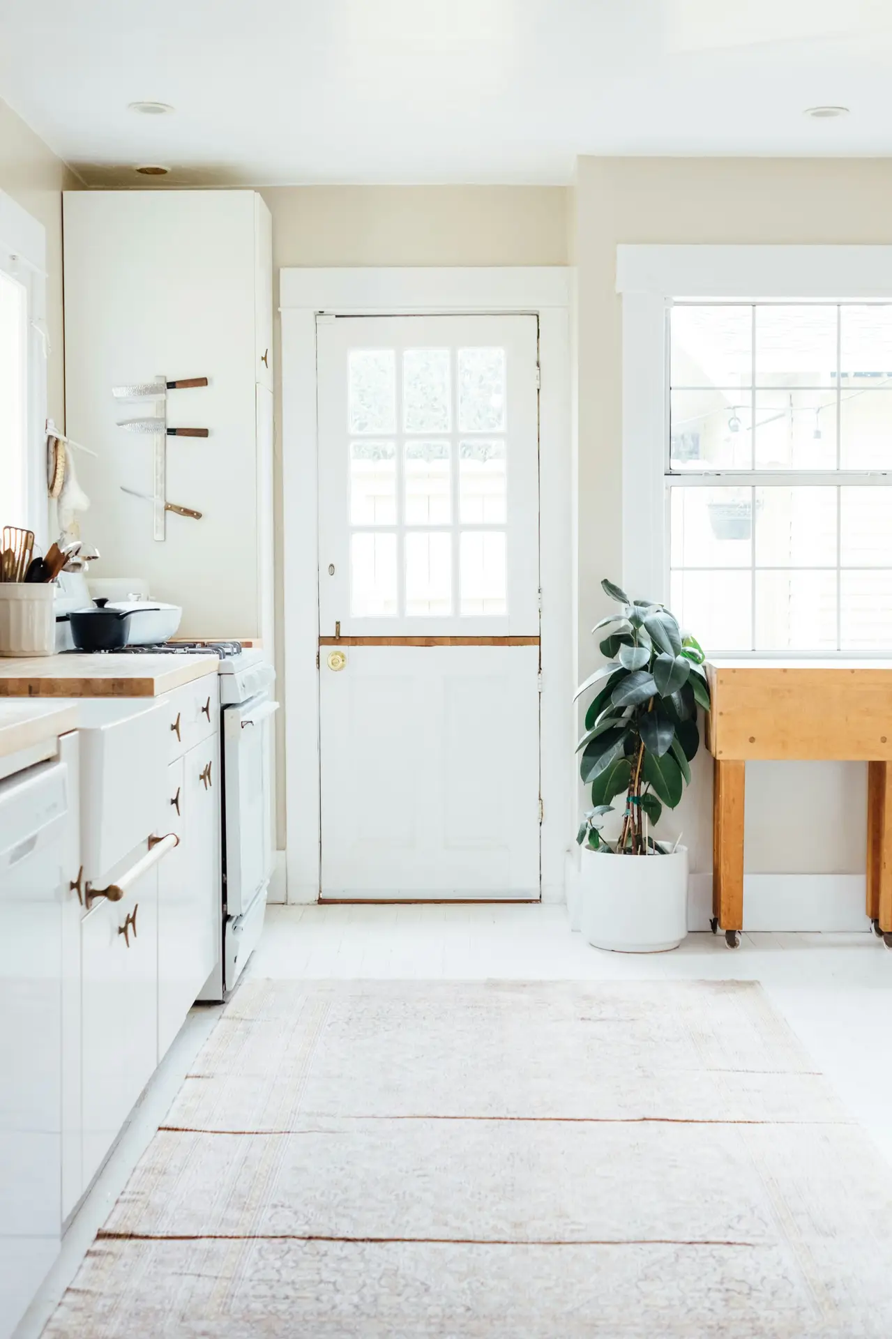 Bright and welcoming view of modern farmhouse kitchens, featuring white cabinetry, warm wood countertops, a glass-paneled door, and a soft vintage rug layered on light floors, complemented by natural light, simple cookware, and a potted plant for a relaxed, homey feel.