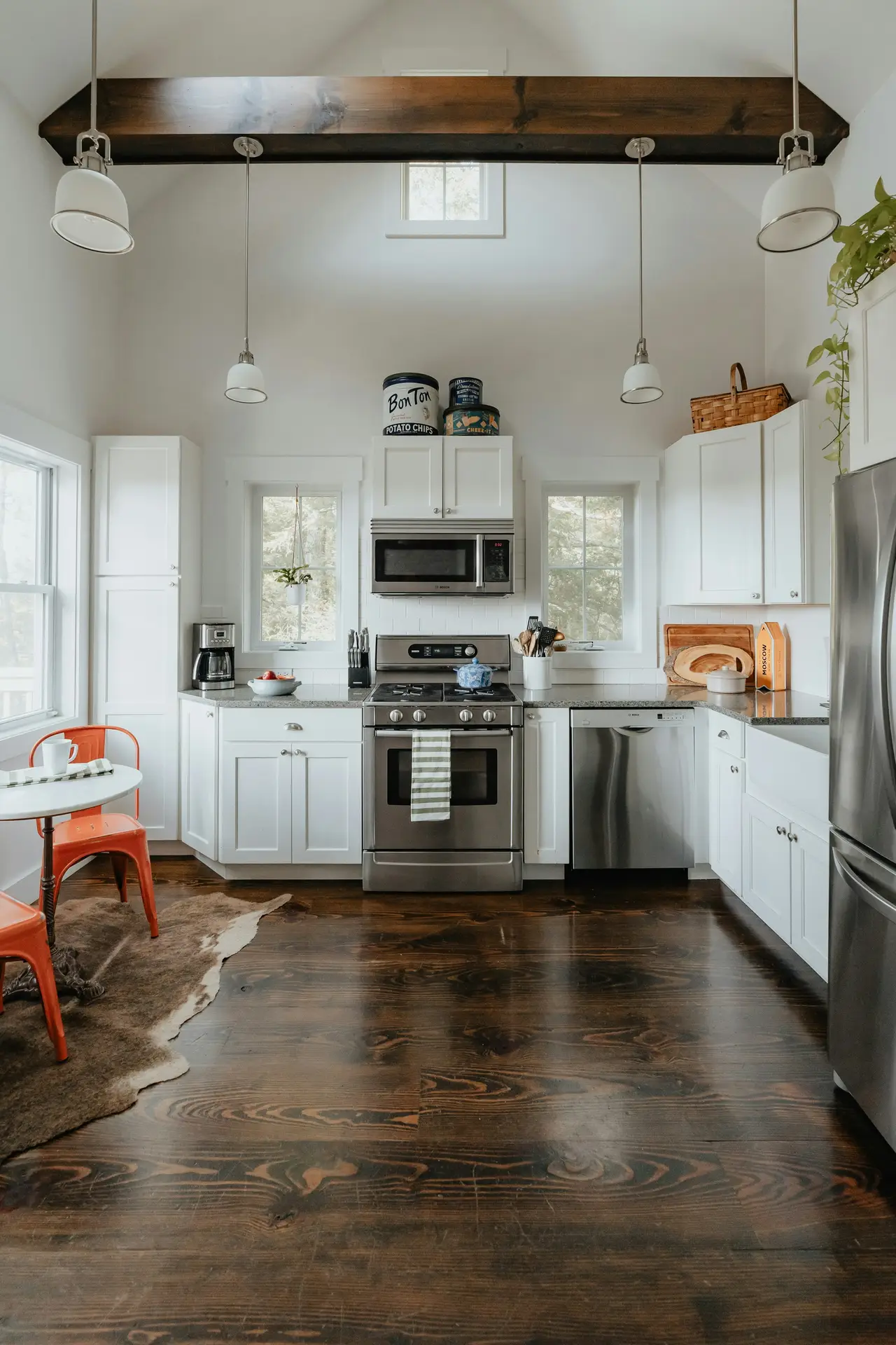 Spacious and inviting view of modern farmhouse kitchens, featuring a wooden beam on the ceiling, crisp white cabinets, stainless steel appliances, dark wood floors, and simple pendant lights, with natural light streaming through multiple windows to create a warm, lived-in atmosphere.