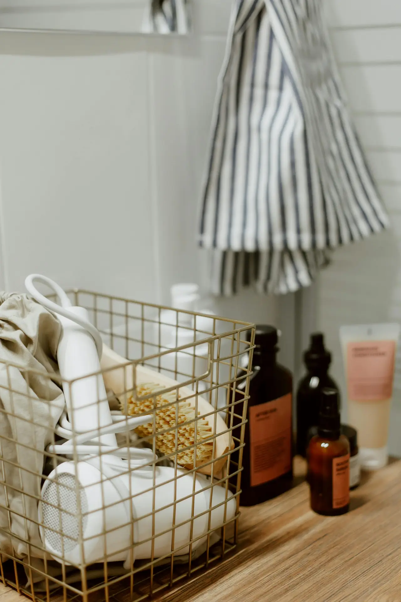 Bathroom countertop styled with practical organization ideas for the bathroom, featuring a gold wire basket used as bins for hair care products holding a white hair dryer, brush, and fabric pouch, alongside amber glass bottles and neatly arranged toiletries on a wooden surface with a striped towel hanging in the background.