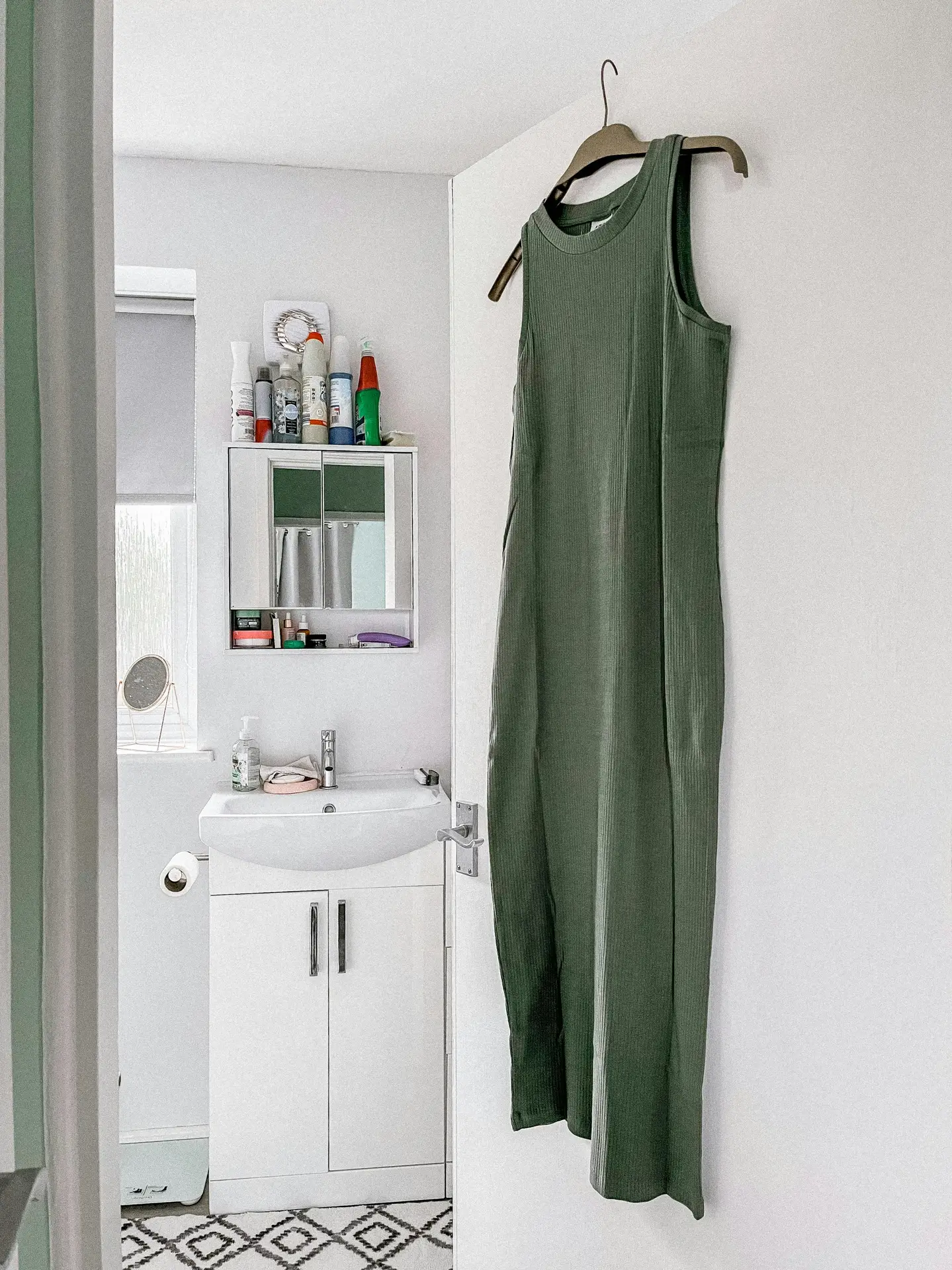 Small white bathroom featuring clever organization ideas for the bathroom, including a clothes hanger on the door holding a long green dress, a compact vanity with storage cabinet, wall-mounted shelf filled with toiletries, and neatly arranged essentials in a bright, minimalist space.