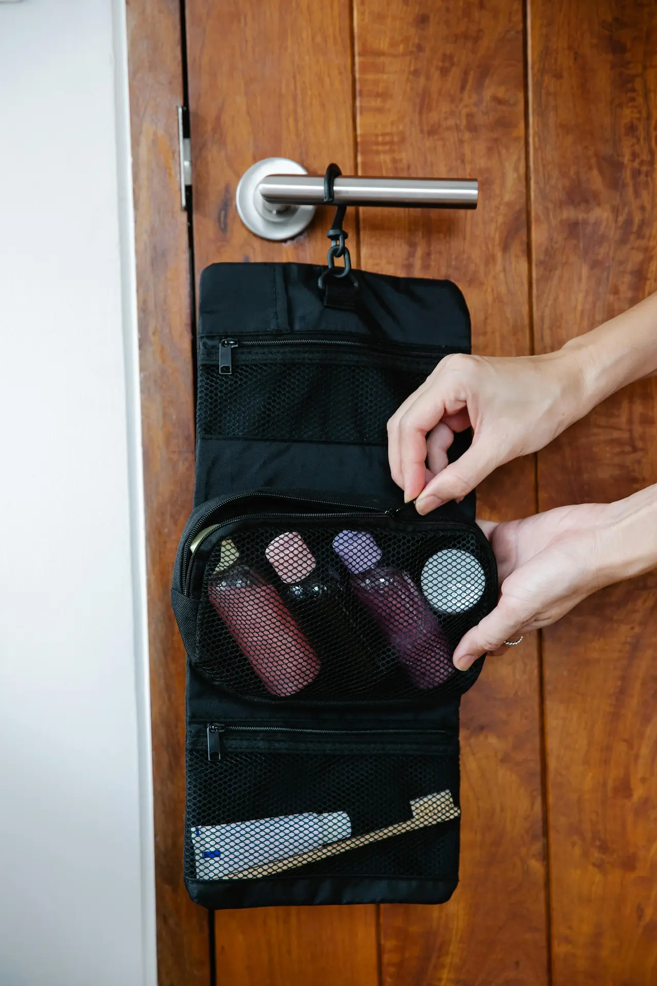Compact hanging toiletry bag hooked over a wooden door handle, with zippered mesh compartments holding small bottles and a toothbrush, while hands adjust the pocket-an example of smart organization ideas for the bathroom that keeps essentials visible, portable, and neatly stored without taking up counter space.
