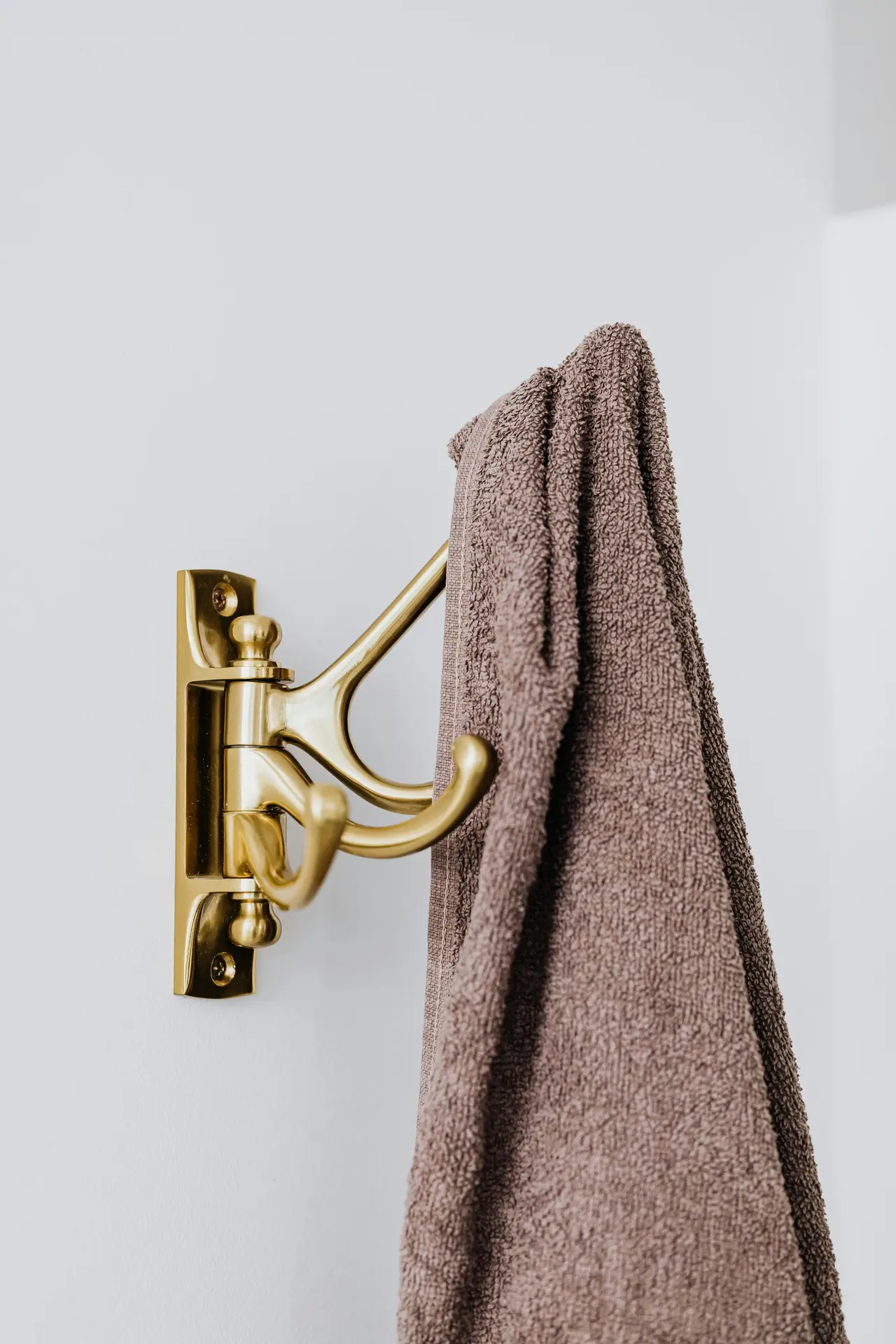 Close-up of a brushed brass wall-mounted double hook holding a soft taupe towel against a light gray wall, showcasing practical organization ideas for the bathroom and stylish hooks for towels that keep linens within easy reach while adding a subtle touch of elegance.