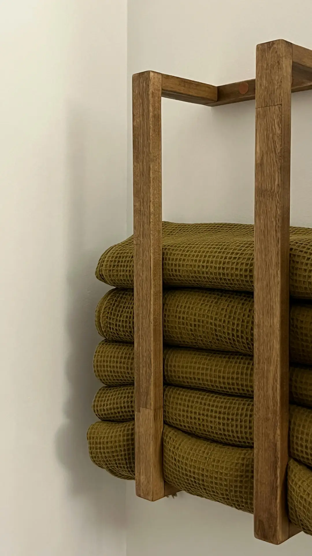 Close-up of a wooden wall-mounted rack neatly stacked with folded olive-green towels, creating a warm and natural accent against a light wall. This setup showcases practical organization ideas for the bathroom, using compact towel holders to keep linens tidy while adding texture and a spa-inspired feel to the space.