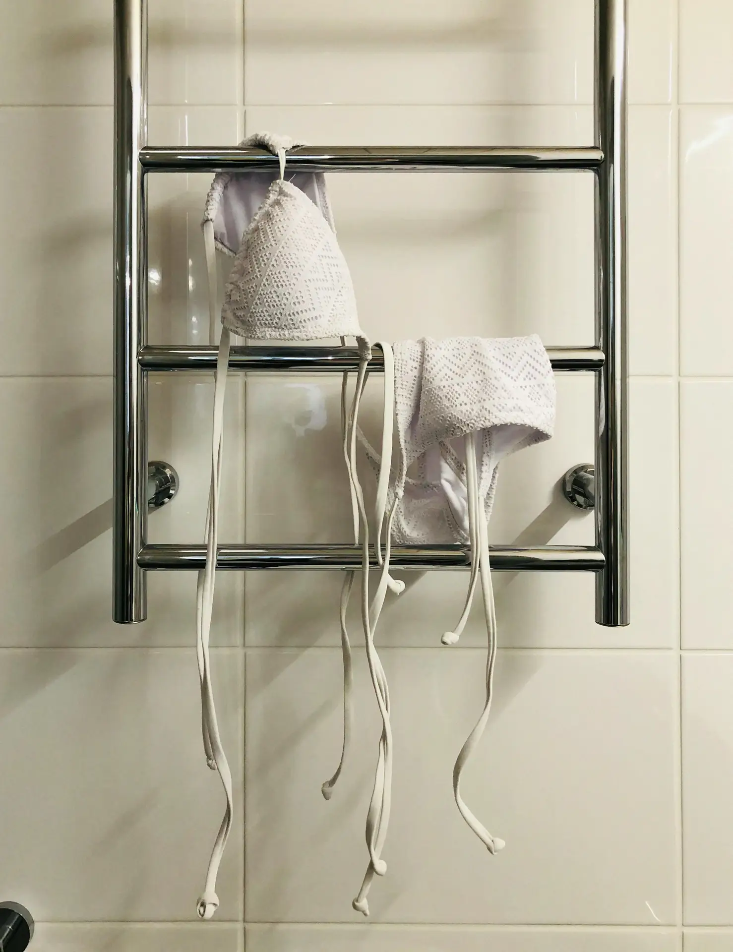 Chrome wall-mounted bars fixed to glossy white tiles hold a white lace bikini top and matching bottoms, neatly draped to dry after use. This setup reflects practical organization ideas for the bathroom, showing how rails for towels and clothes can double as a simple drying solution while keeping the space tidy and uncluttered.