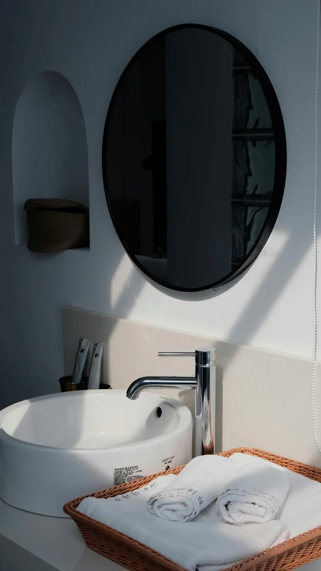 Minimalist bathroom scene with a round black-framed mirror above a white vessel sink and sleek chrome faucet. Soft sunlight casts shadows across the wall, while neatly rolled white linens rest in a woven tray on the counter-an elegant example of organization ideas for the bathroom that shows how to store guest towels in small baskets for a tidy, welcoming touch.